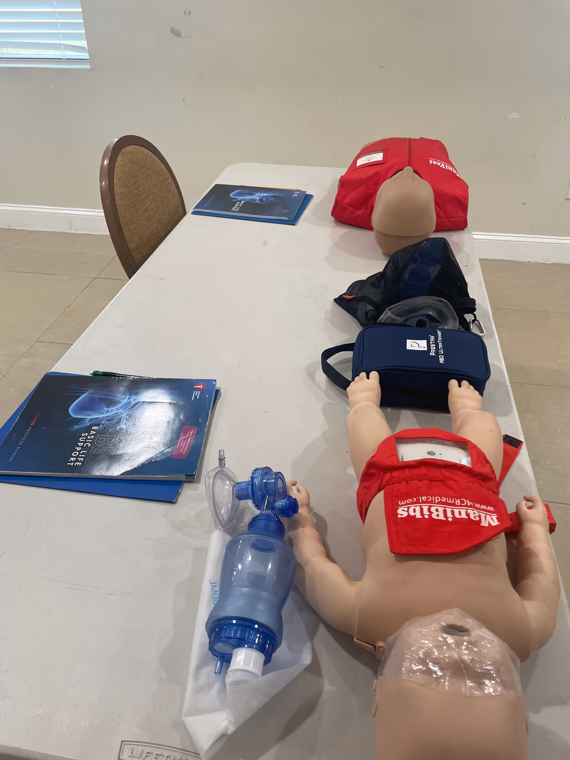 CPR training mannequin on a table with resuscitation mask, bag, and manuals in a room