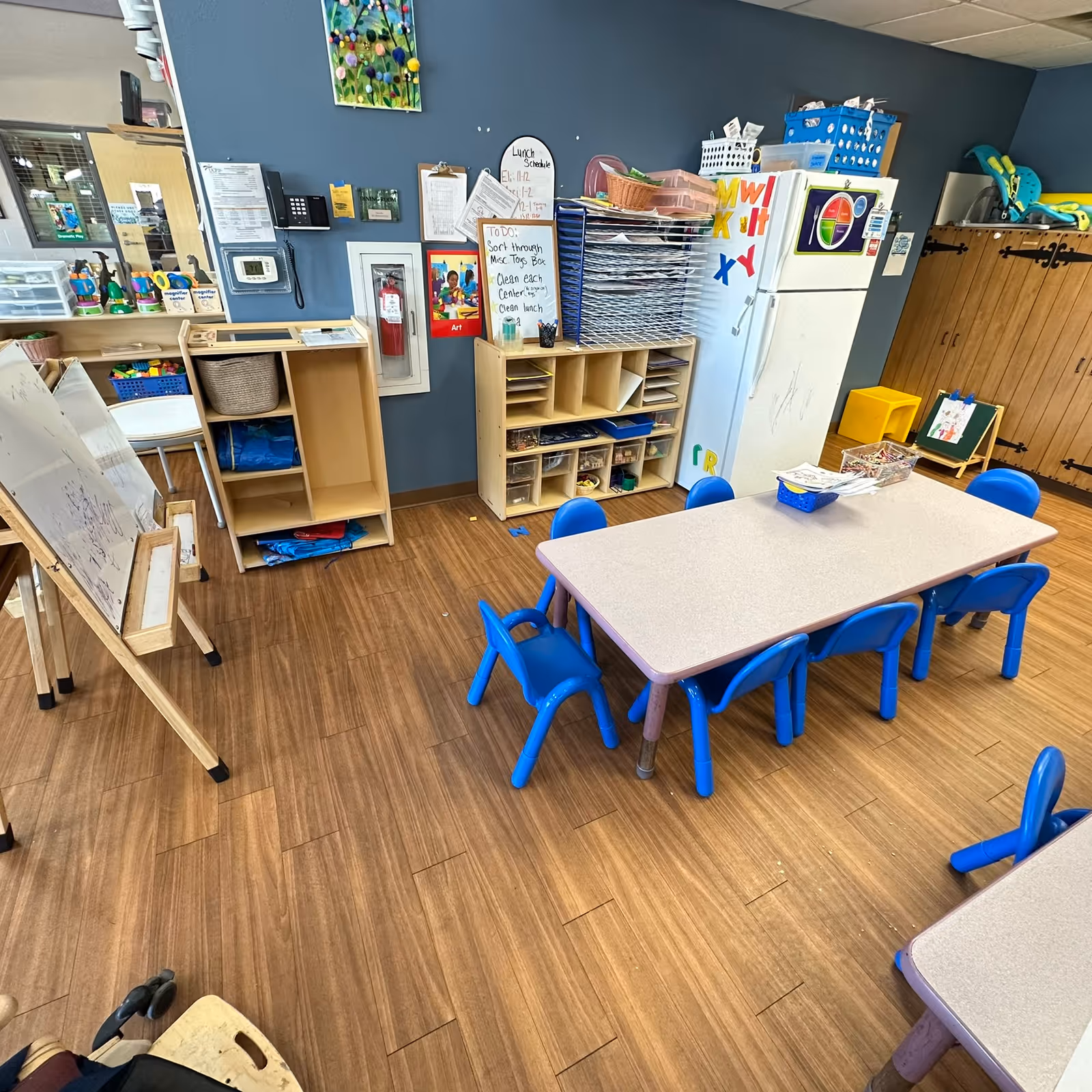 Colorful classroom-like activity room with tables, blue chairs, storage cubbies, and art supplies.