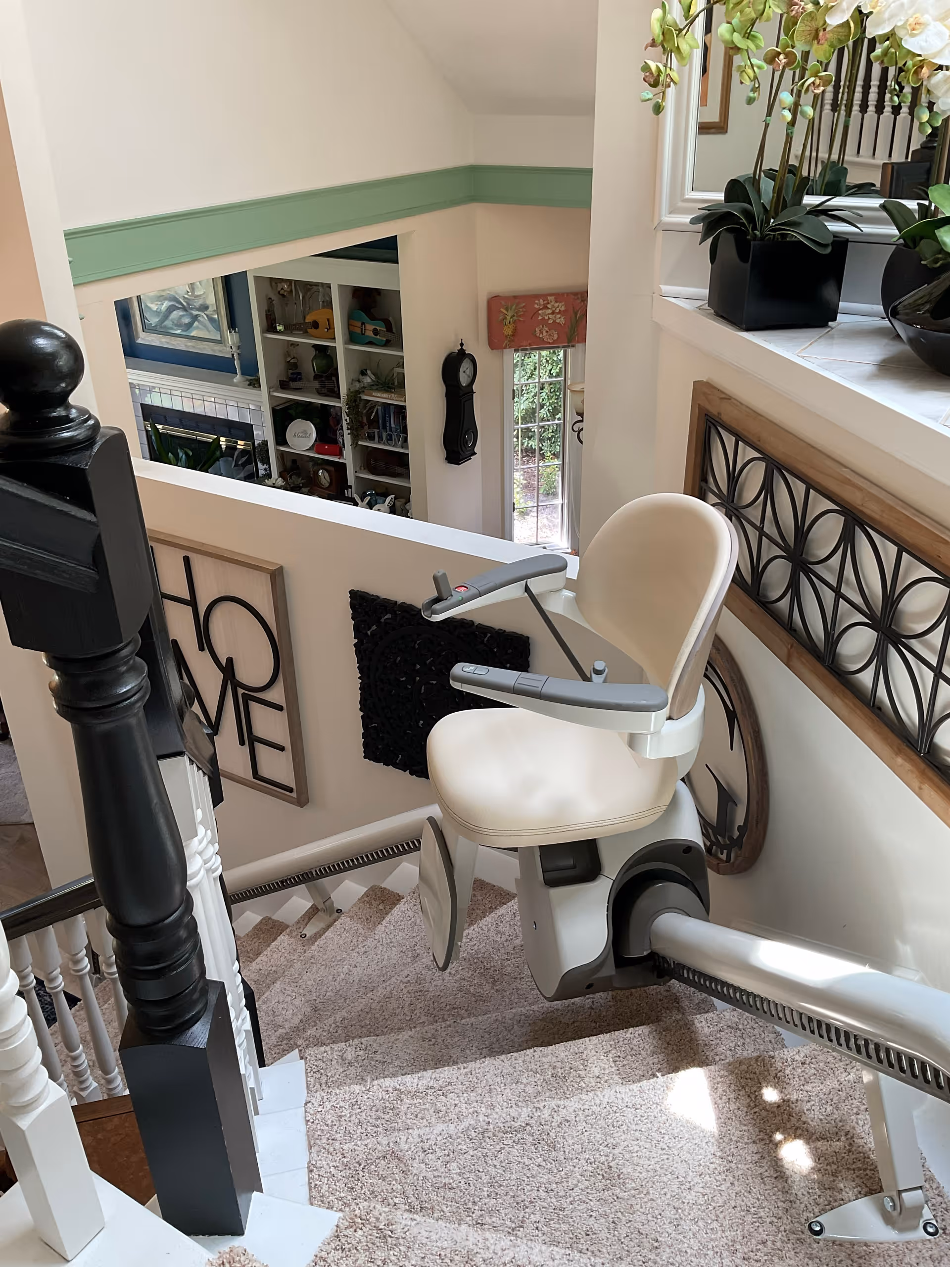 Interior staircase with beige stairlift chair, decorative railing, and potted plants nearby.