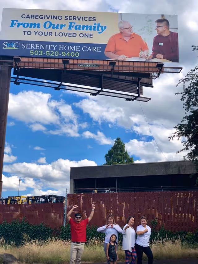 Large outdoor billboard atop a pole promoting Serenity Home Care, with people posing below.