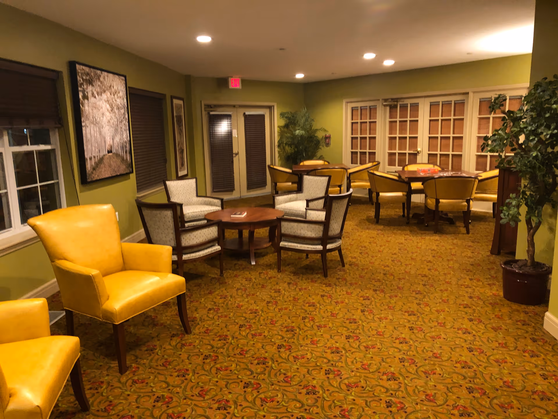 Interior lounge area with yellow chairs, round tables, and green walls in a senior living facility.