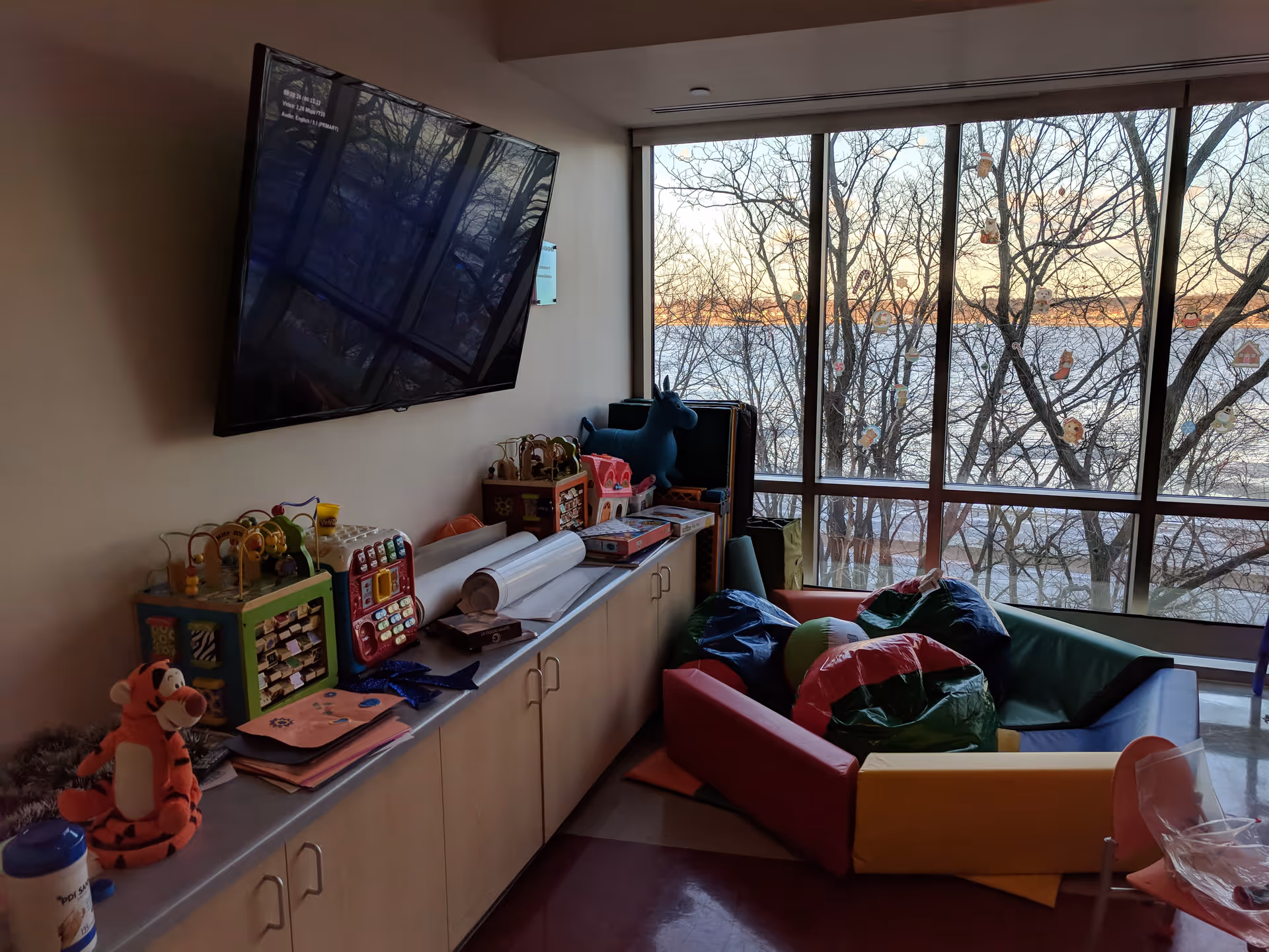 Playroom area with toy shelves, colorful cushions, a wall-mounted TV, and large window.
