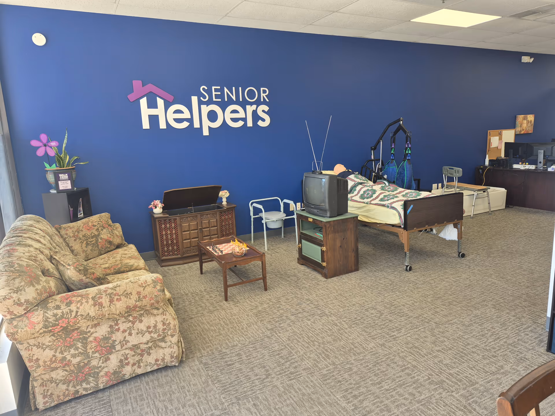 A Senior Helpers interior living area with a bed, couch, TV, and blue wall sign.