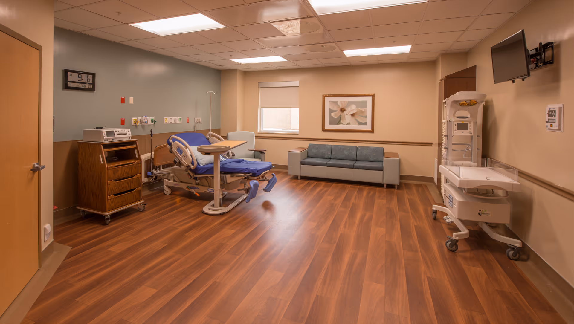 Hospital-style patient room with adjustable bed, medical equipment, seating, and wall-mounted monitor.
