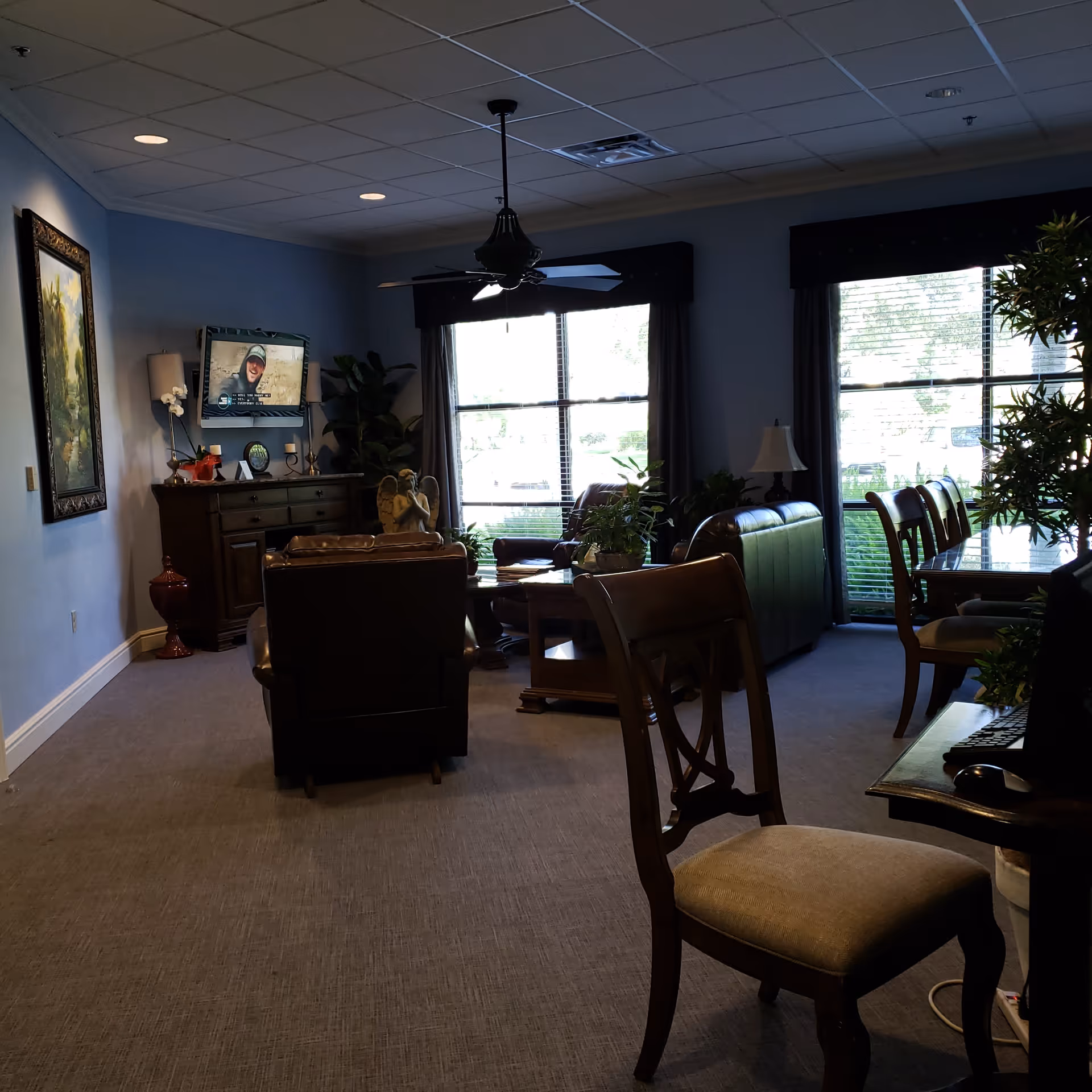 Dimly lit living room lounge with chairs, a sofa, TV, and plants by large windows.