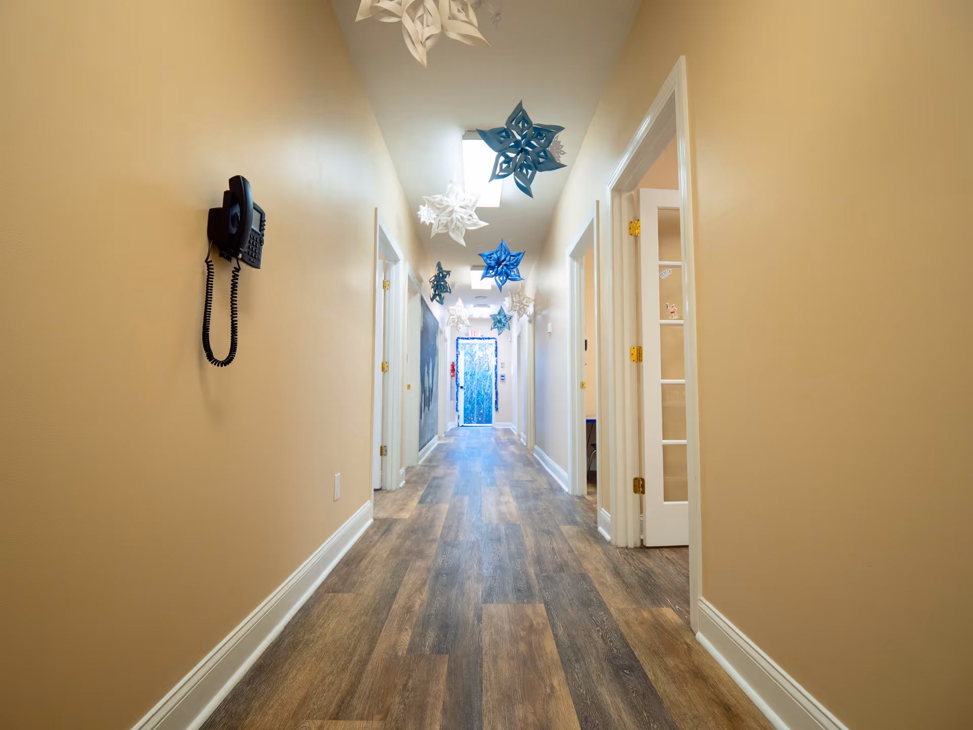 A long beige hallway with doors on both sides, wooden floor, and hanging snowflake decorations.
