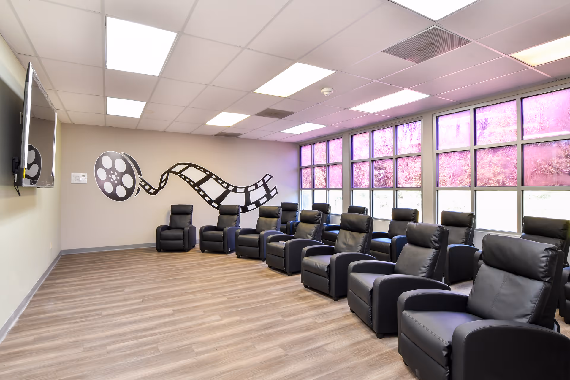 Spacious room with rows of black leather recliners facing a film reel mural and large windows.