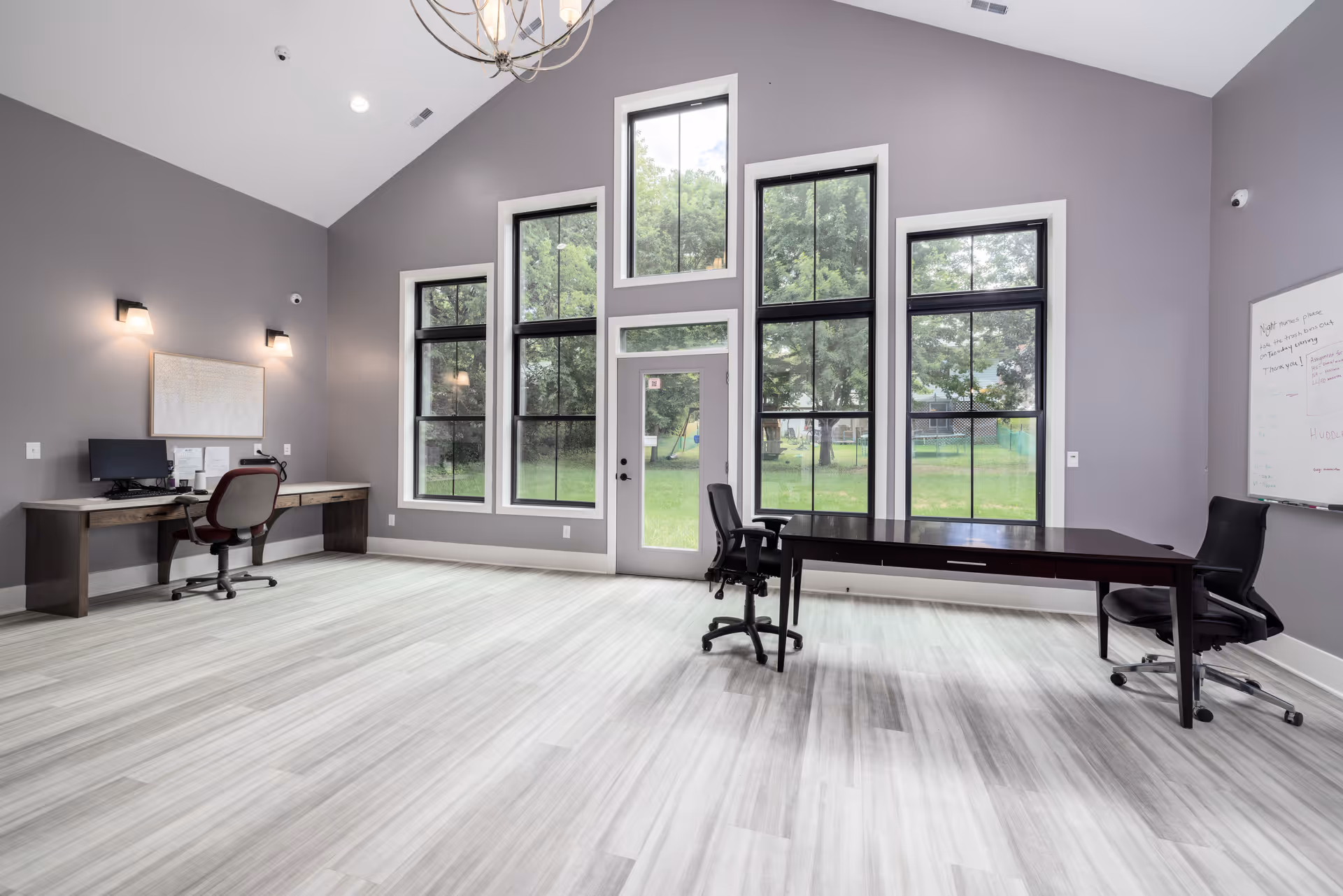 Bright interior room featuring desks, chairs, a whiteboard, and large windows overlooking a green lawn.