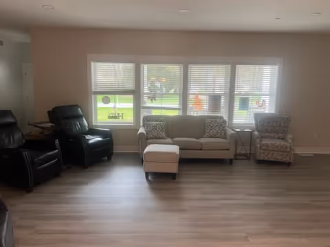 Bright living room with sofa, ottoman, and multiple chairs by large windows.