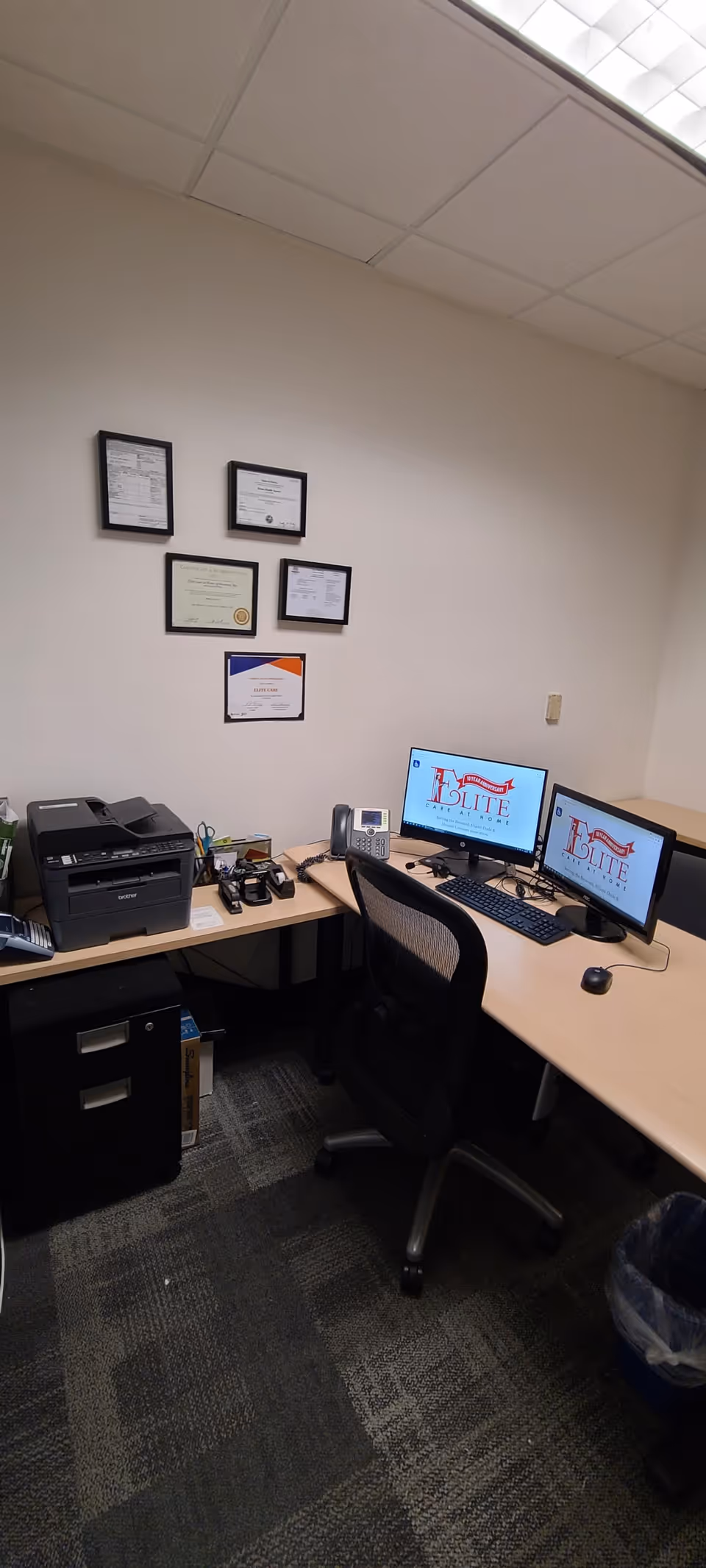 Office workspace with dual computer monitors, printer, and framed certificates on the wall.