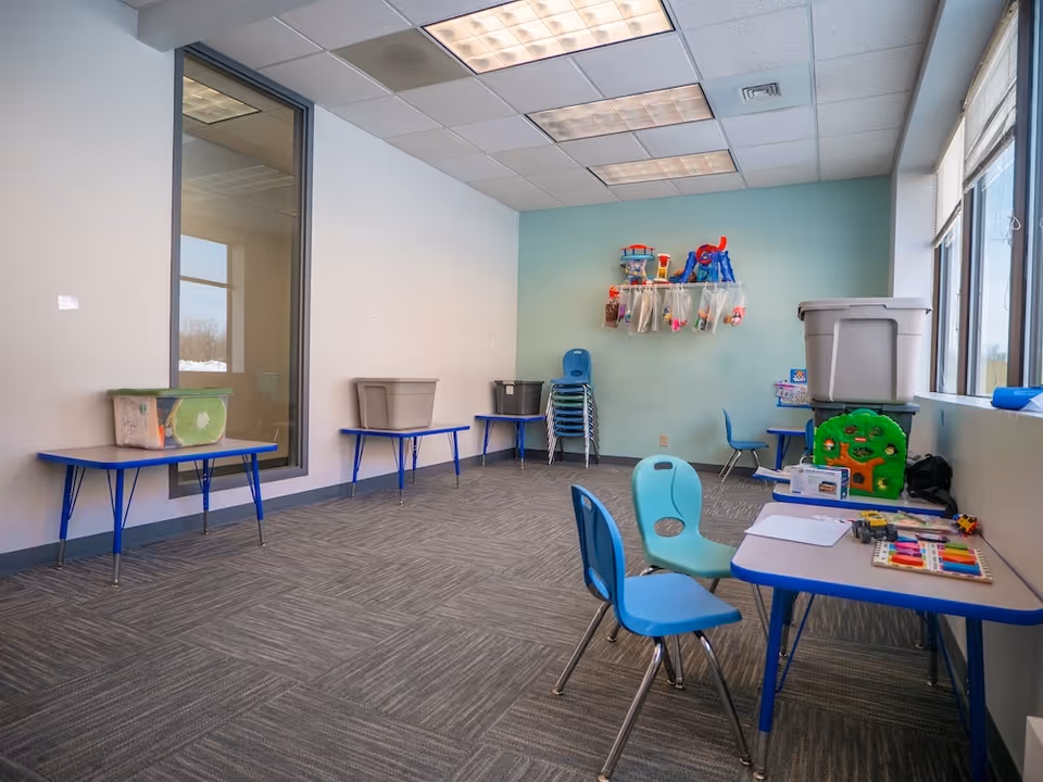 Bright classroom-like room with small blue chairs, tables, shelves, and colorful toys along the wall.
