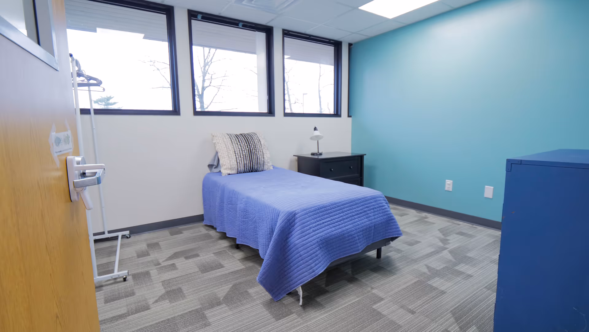 Bedroom in a senior living facility with a blue bedspread, pillow, and a nightstand near windows.