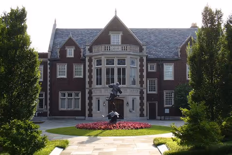 Brick-fronted stately building with statue, circular flower bed, and tall trees.
