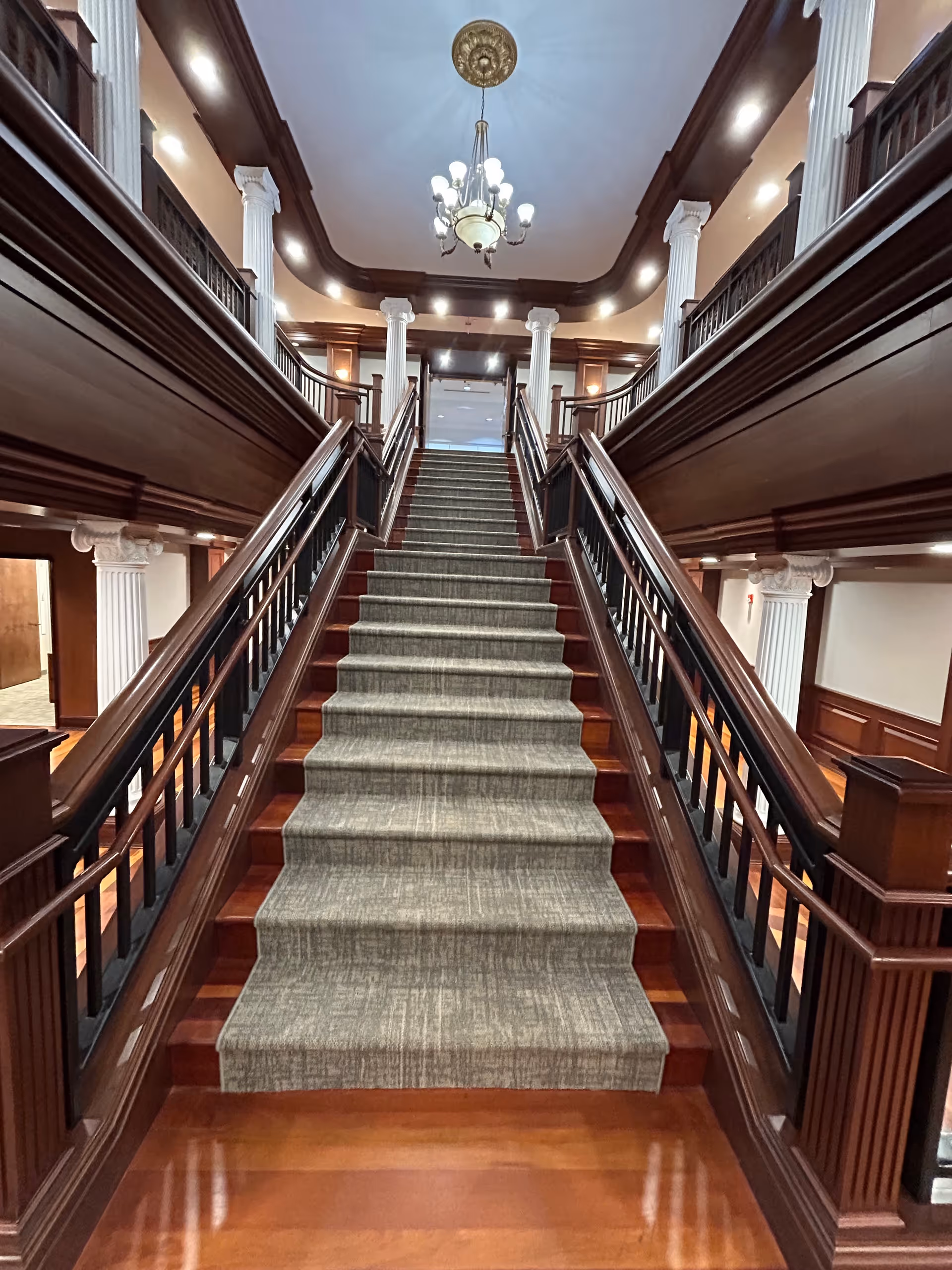 Wide wood staircase with carpeted steps, railings, columns, and chandelier in a grand interior.