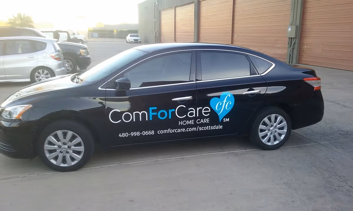 Black ComForCare Home Care sedan parked in a paved lot beside warehouse doors.