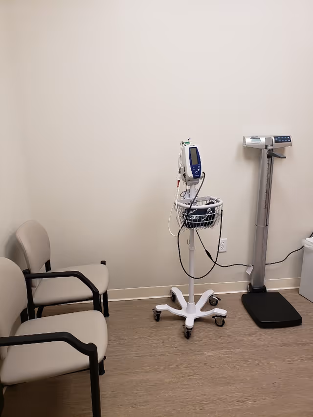 A small medical examination room with a wheeled monitor, scale, and two chairs.