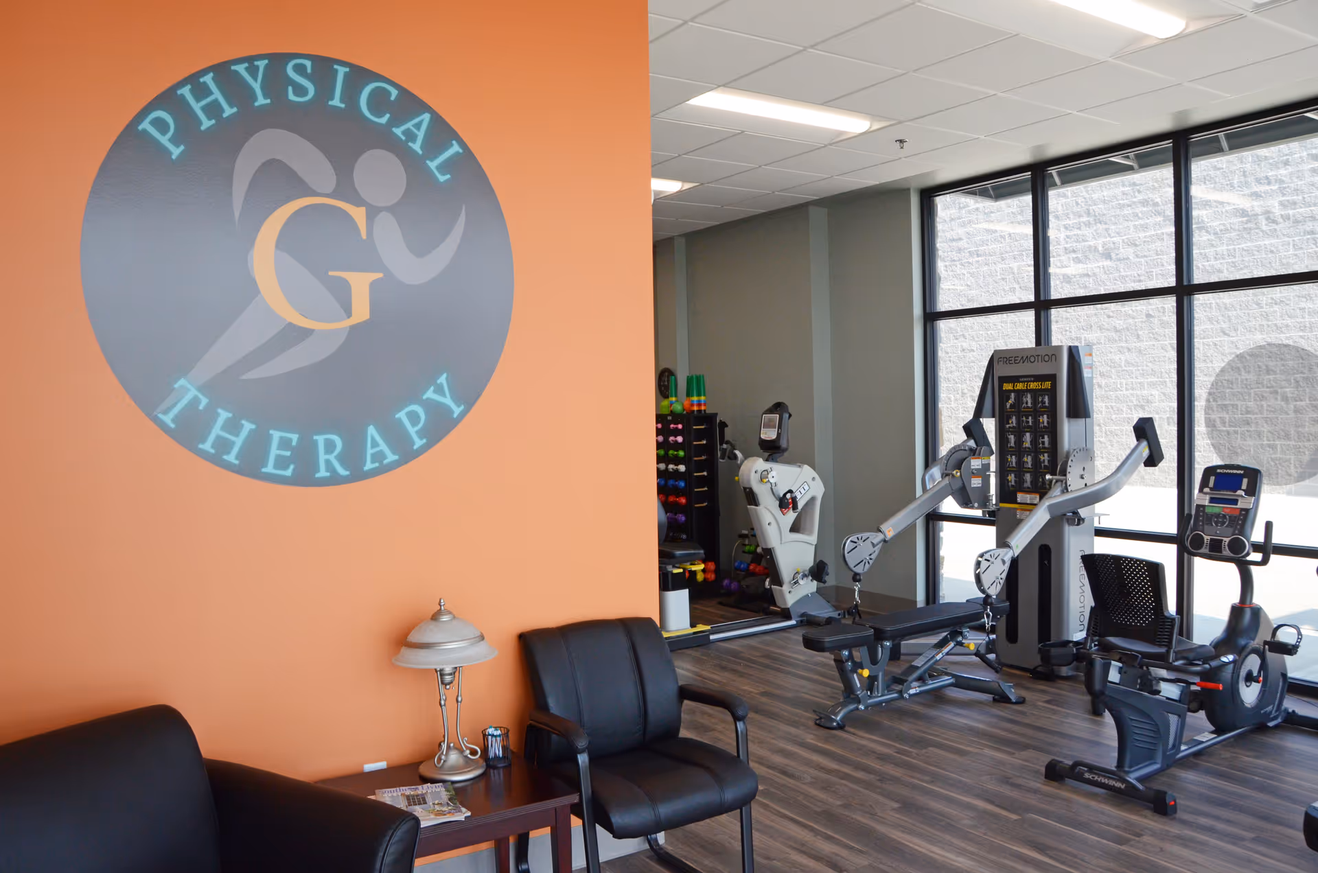 Interior gym area with orange wall logo reading Physical Therapy and exercise machines.