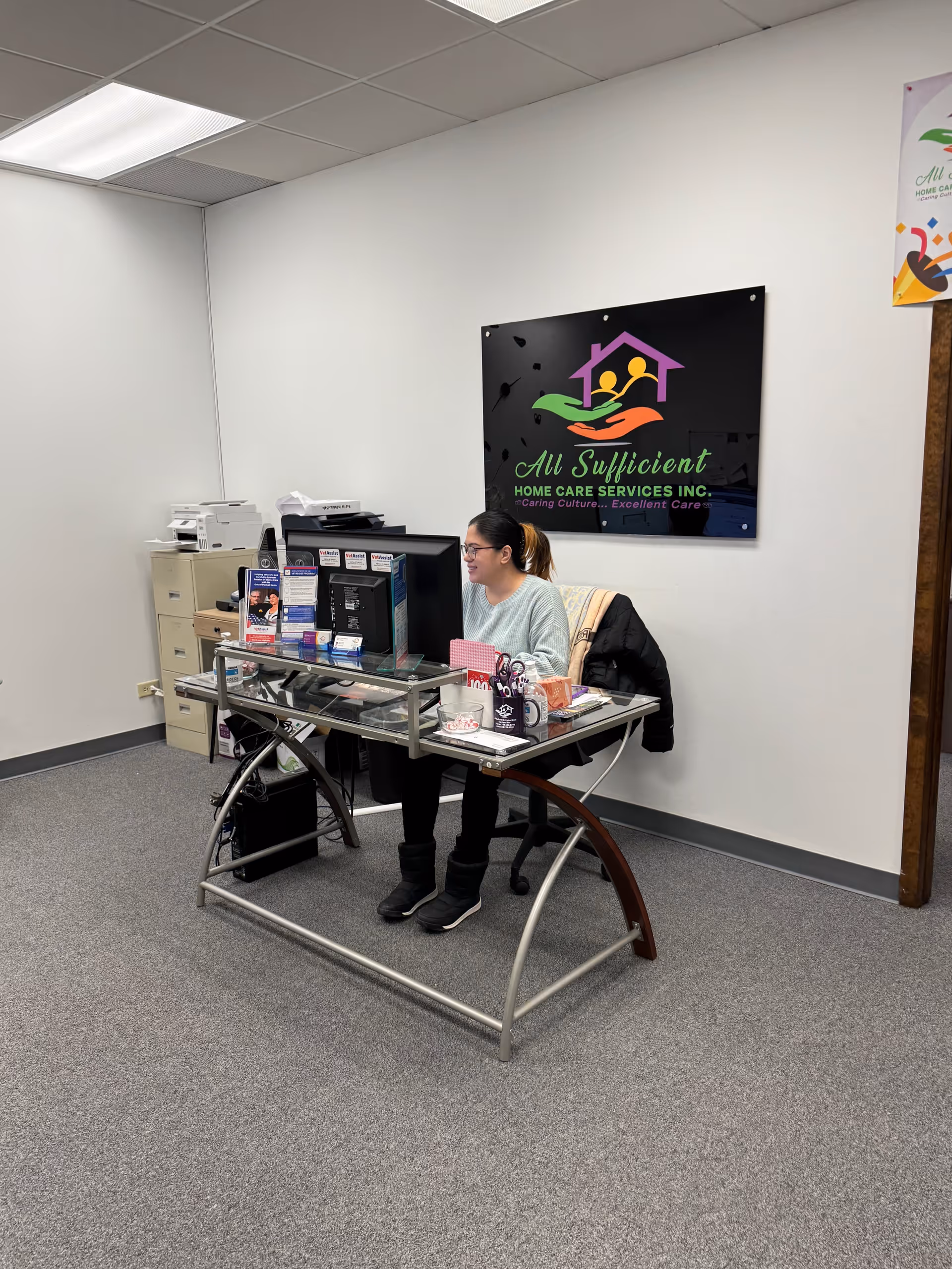 Receptionist sits at glass desk in an office with All Sufficient logo on the wall.