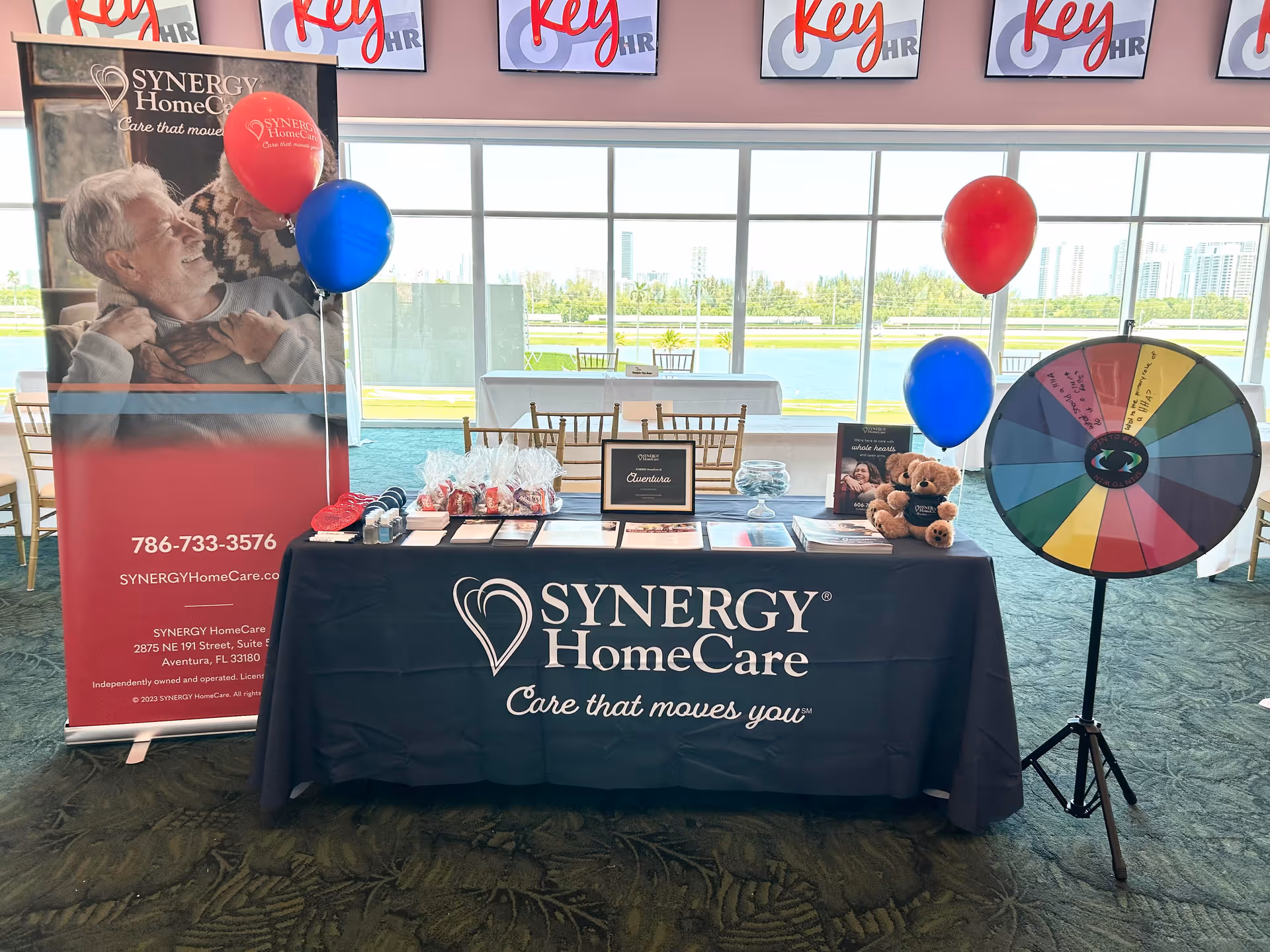 Promotional table for Synergy HomeCare with banners, balloons, and a spinning prize wheel indoors.