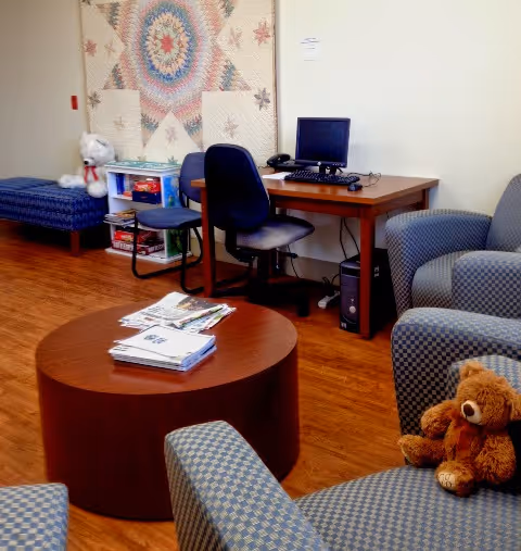 A cozy senior living lounge with a computer desk, round coffee table, and a teddy bear.