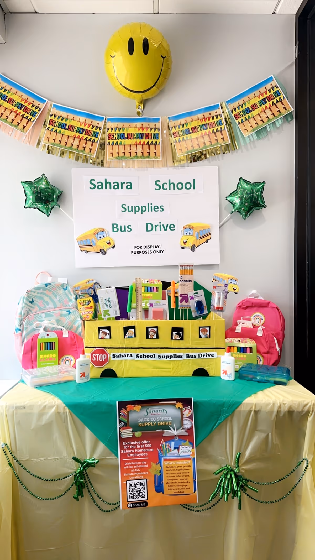 Indoor display of Sahara School Supplies Bus Drive with backpacks, signs, and a yellow smiley balloon.