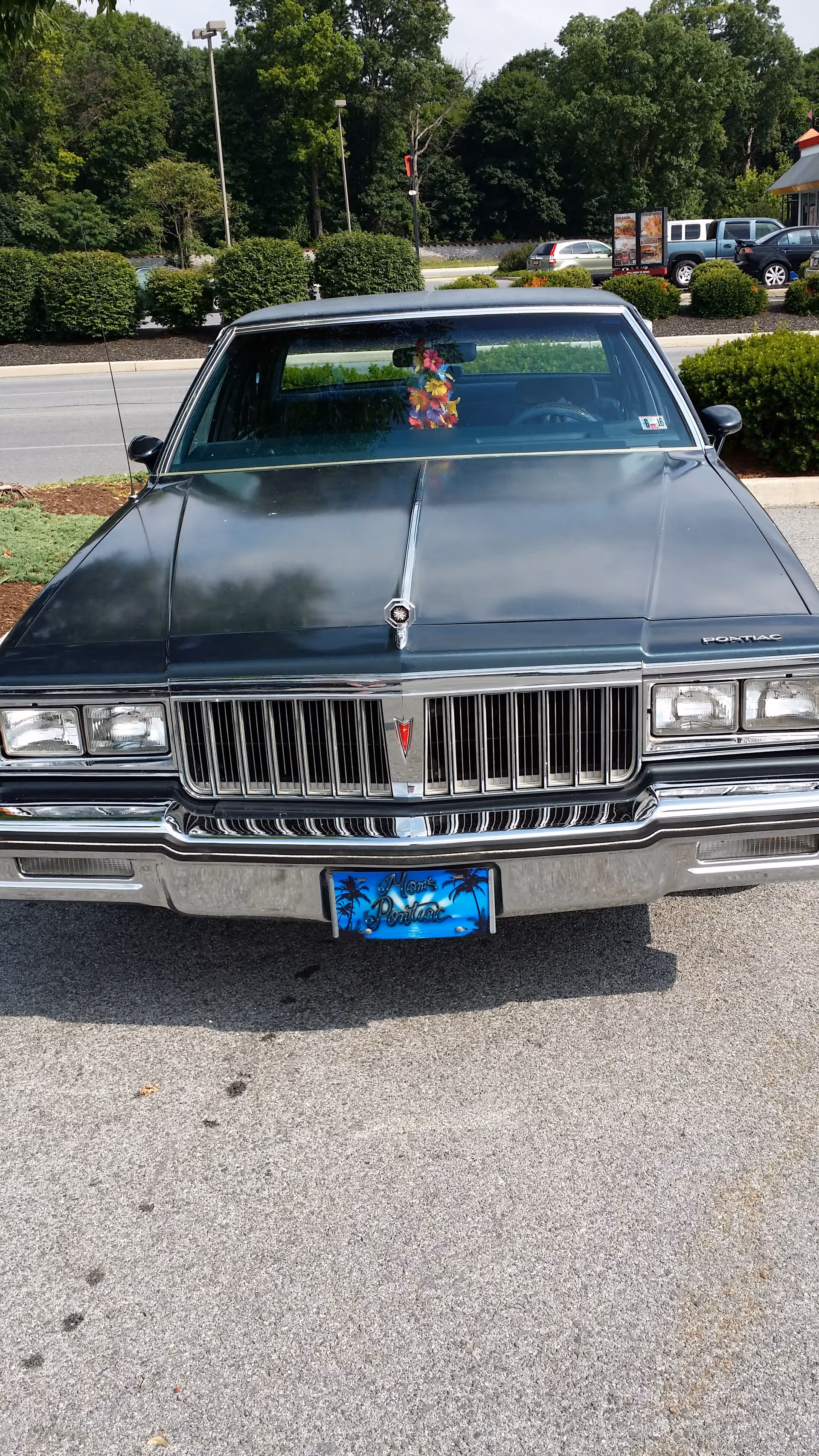 Front view of a dark vintage car parked in a lot with greenery.