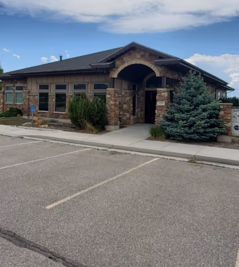 Single-story stone building with arched entrance, shrubs, and a parking lot in front.