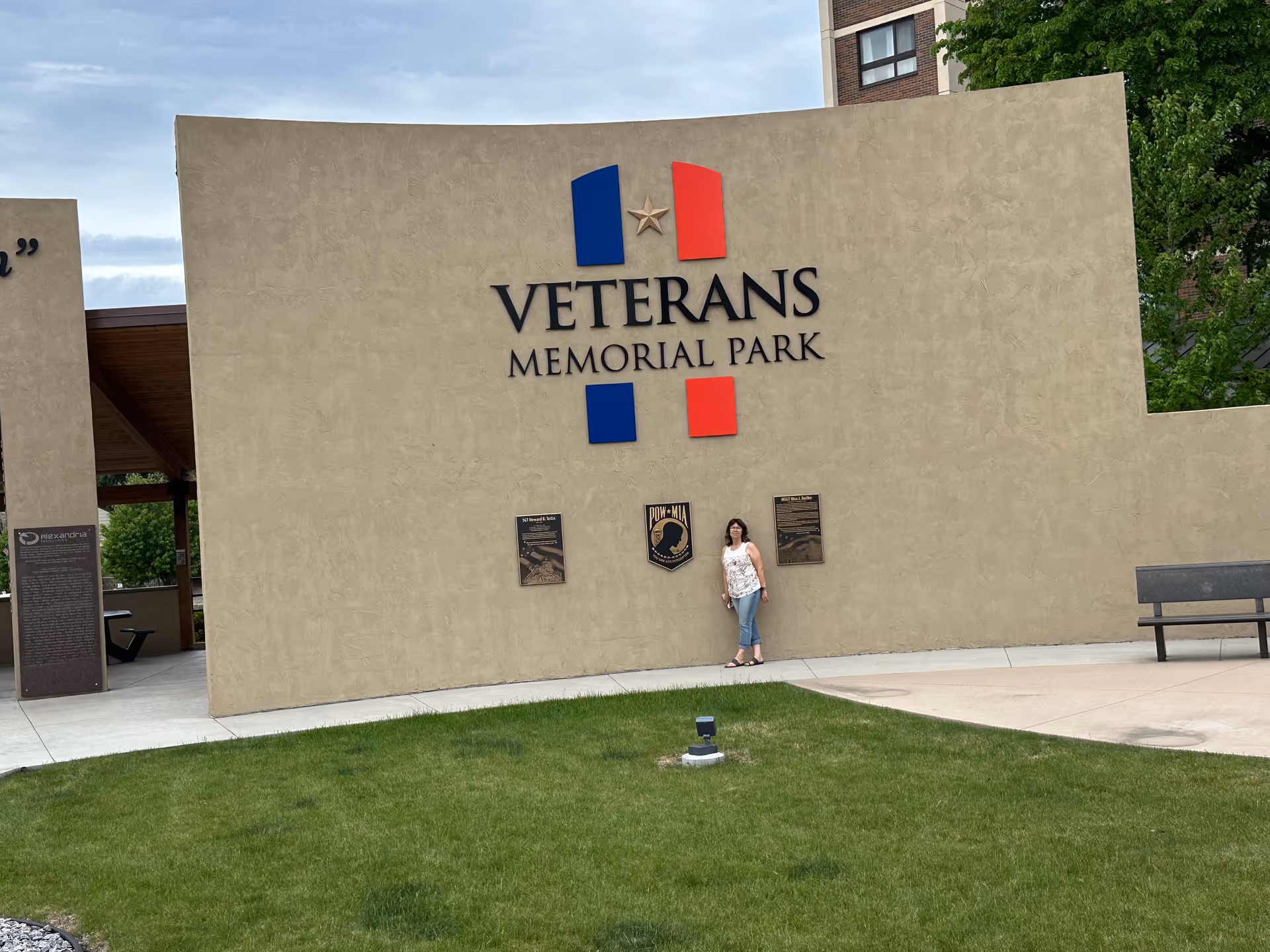 A person stands beside a large Veterans Memorial Park wall with plaques and lawn.