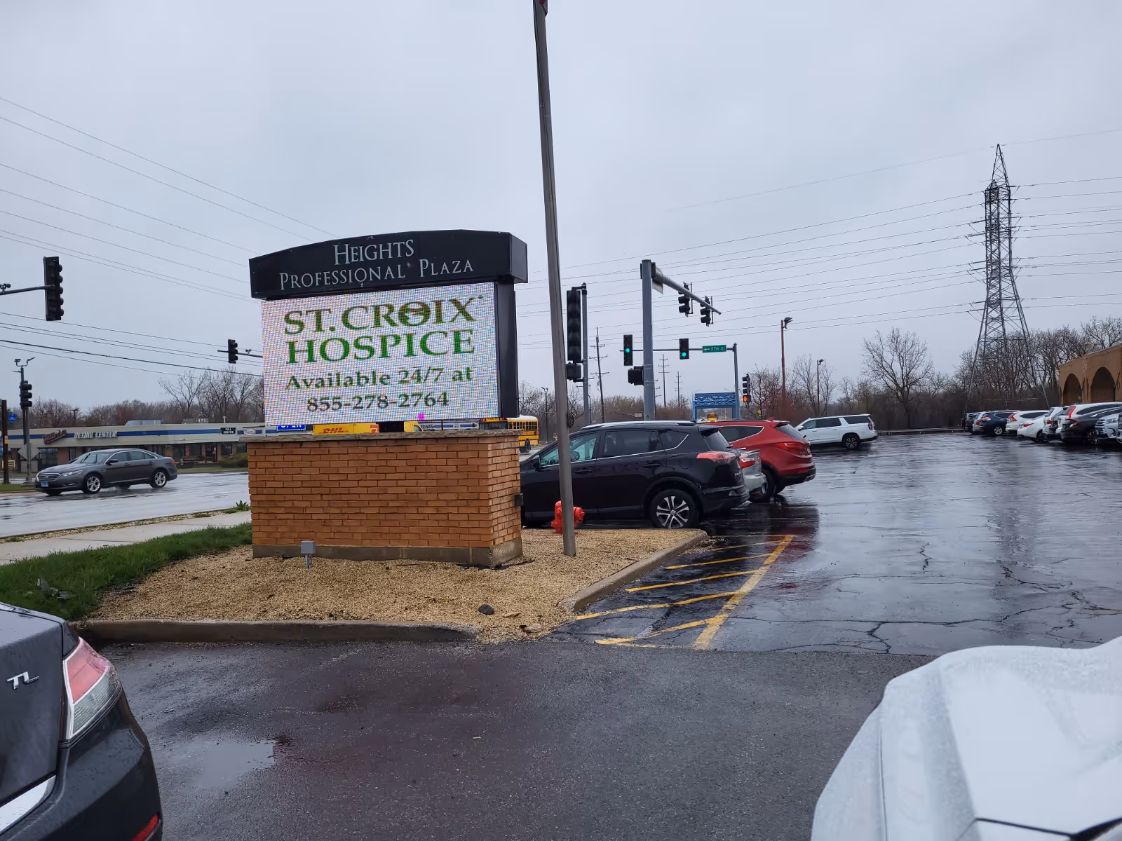 Outdoor parking lot with a St. Croix Hospice sign at a plaza, wet pavement after rain.