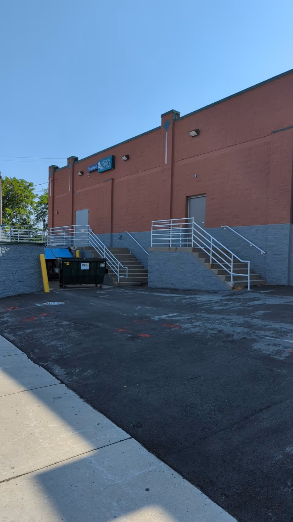 Exterior view of a brick building with staircases, a dumpster, and a paved parking area.
