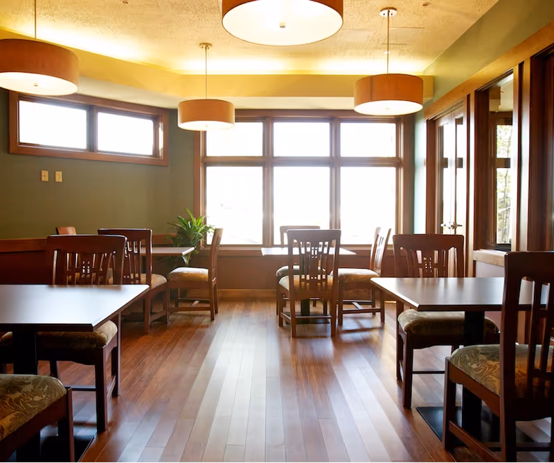 Sunlit dining room with wooden tables and chairs along a windowed wall.