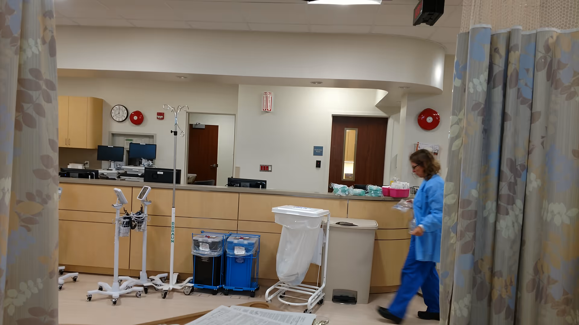 Nurse in blue scrubs walks past a long wooden reception counter inside a medical facility.