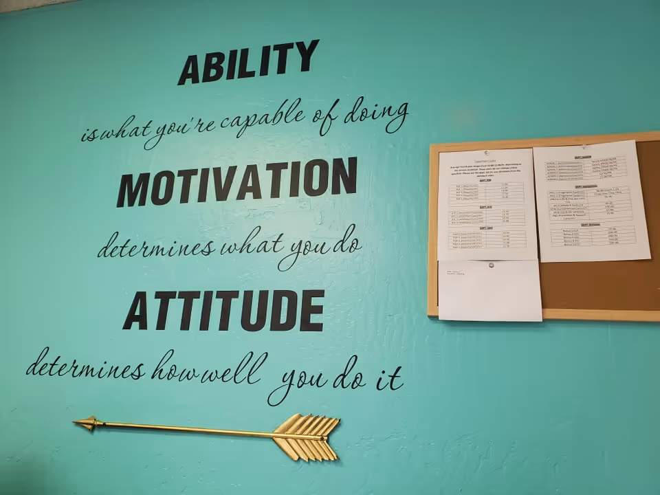 Teal wall displays motivational quote, bulletin board with papers and arrow decoration inside a facility.