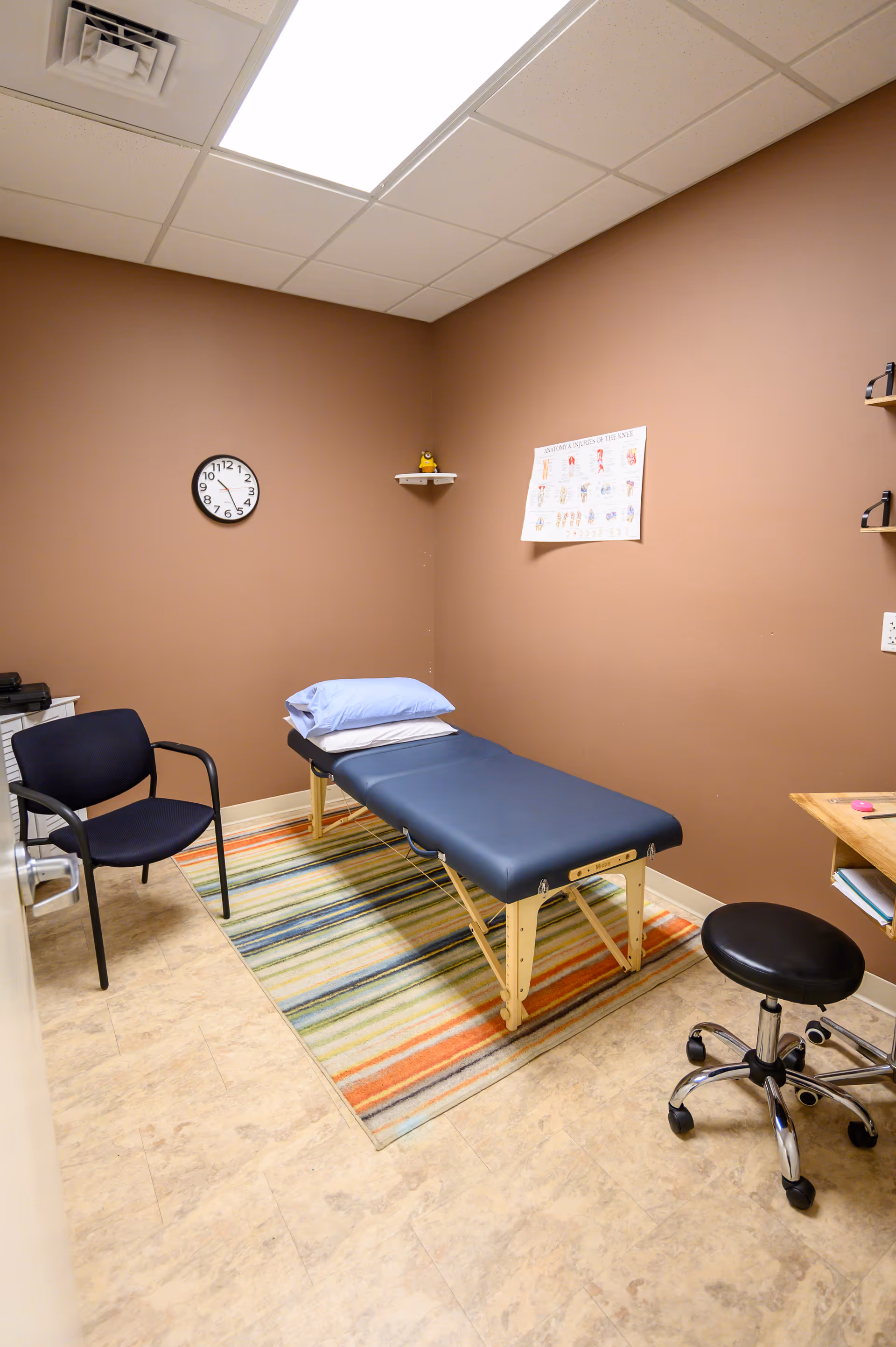 Small therapy room with a blue padded treatment table, chair, stool, and wall chart.