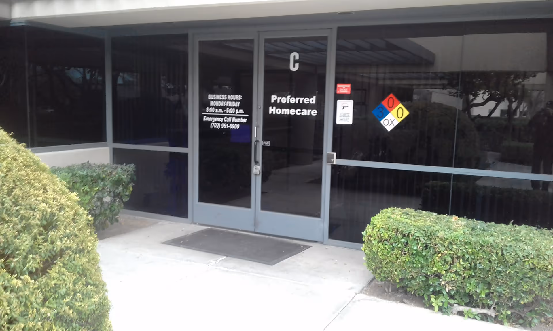 Front entrance of Preferred Homecare with glass doors, business hours sign, and trimmed hedges.
