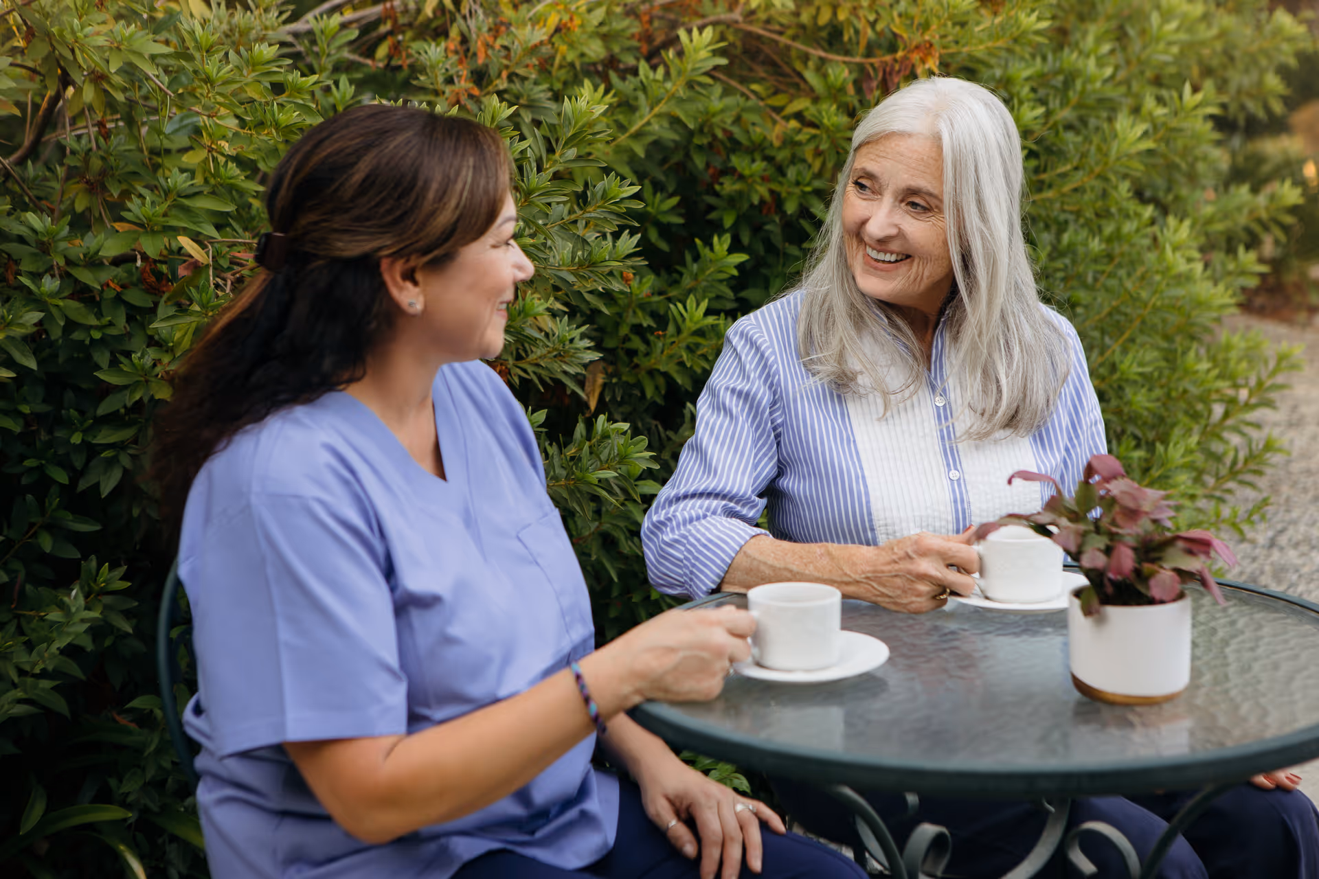 Caregiver in purple scrubs chats with an elderly woman at an outdoor glass table.