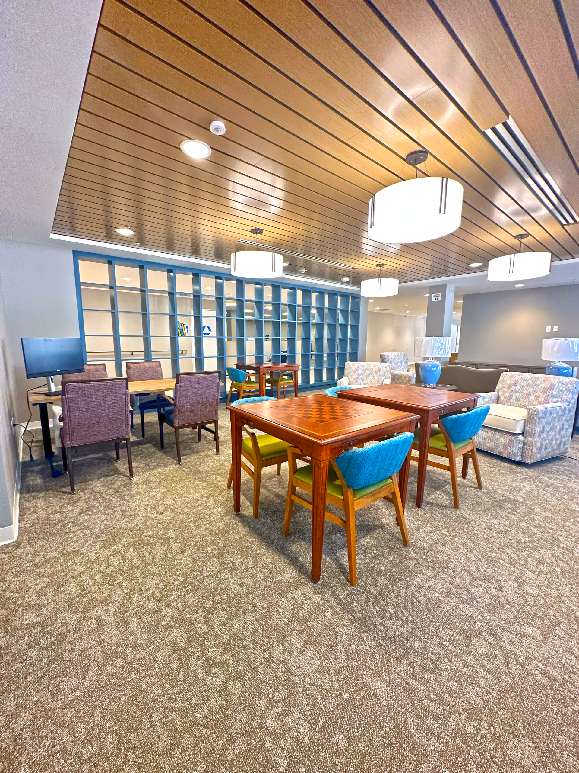 Spacious senior living lounge with tables, chairs, and cushioned seating under a wood-paneled ceiling.