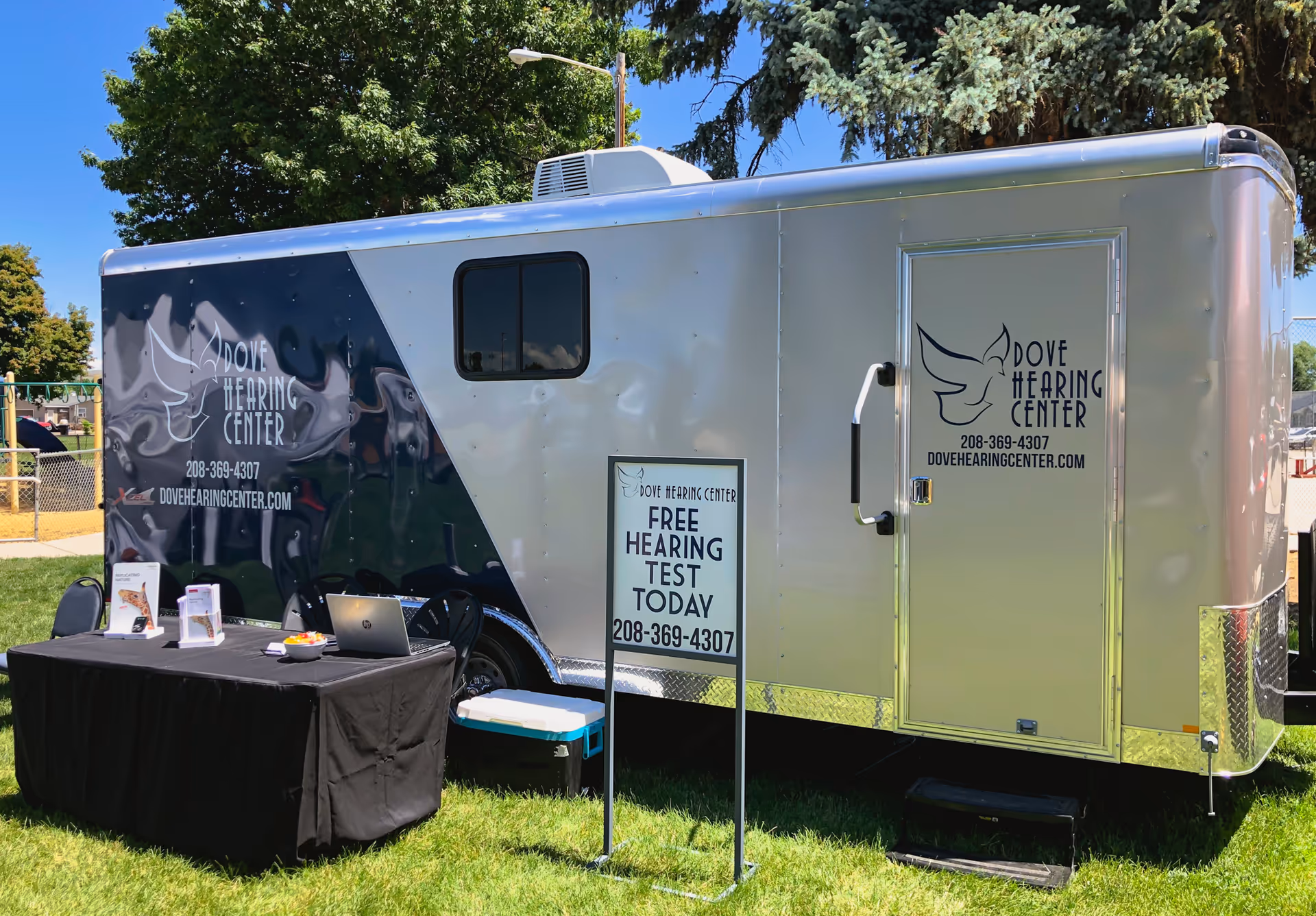 A Dove Hearing Center trailer parked on grass with a table and sign outside.