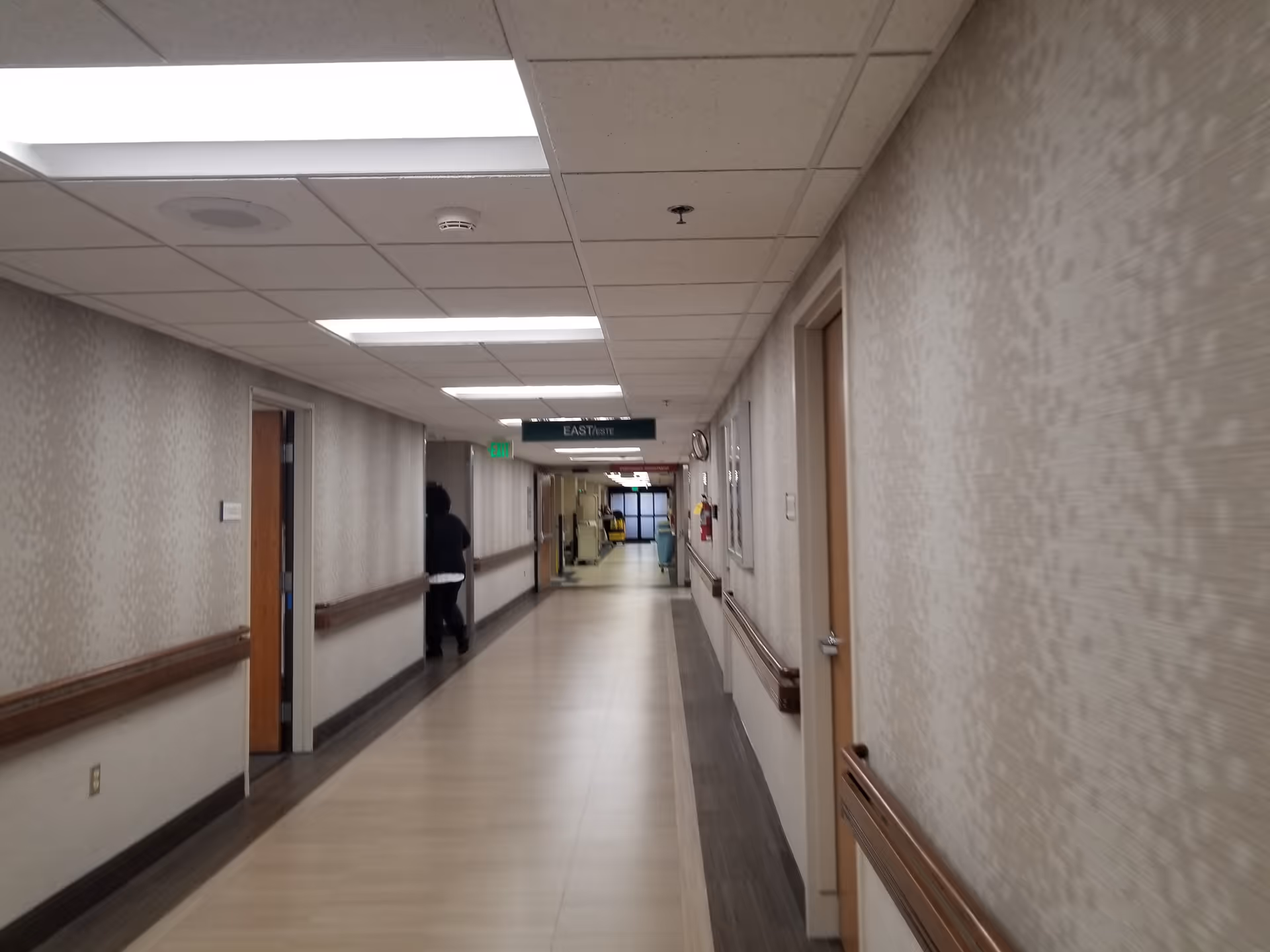 Long, lit hospital corridor with doors on both sides and a person walking away.
