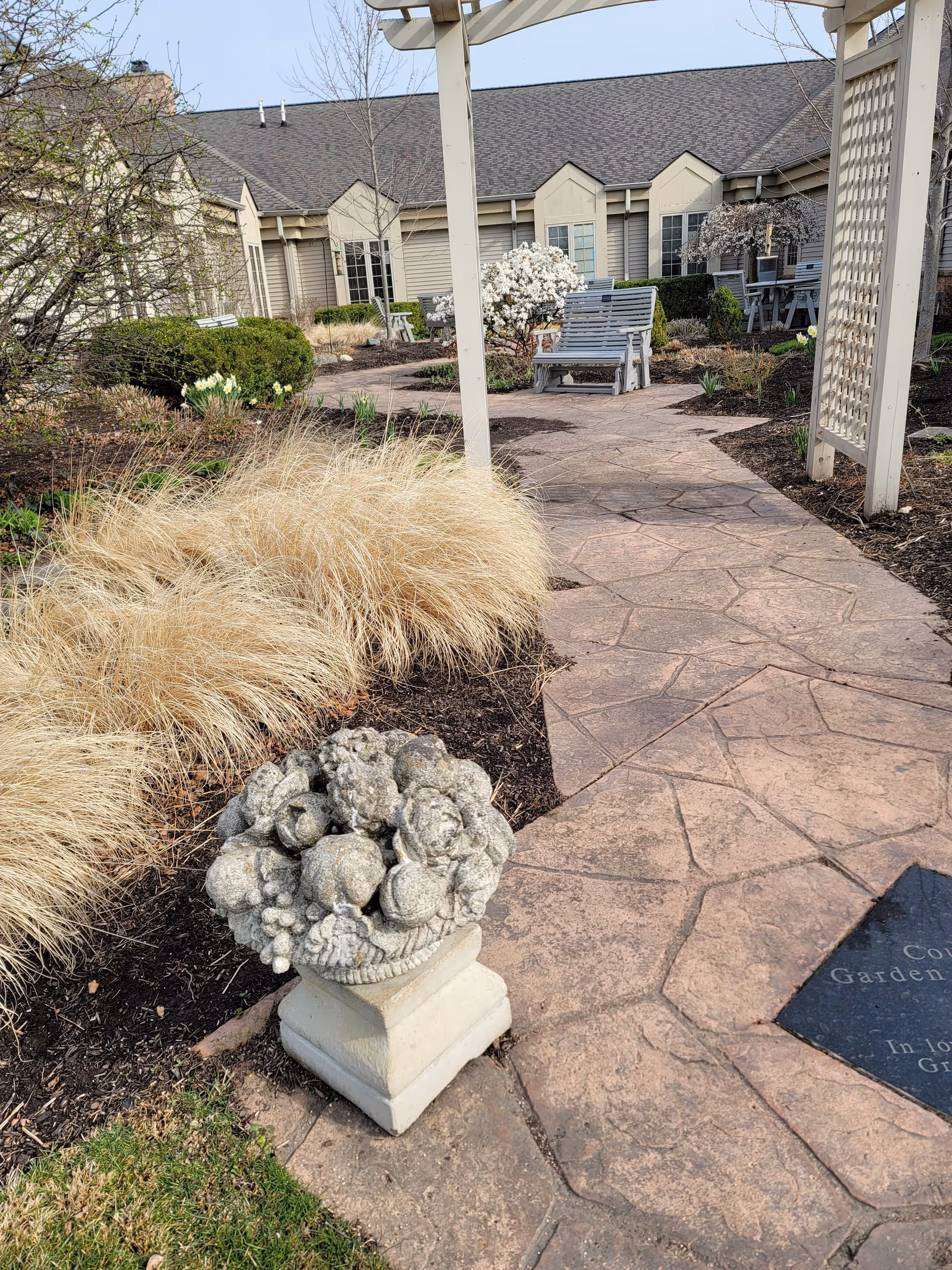 Stone pathway winds through a garden with benches and a pergola near a pastel building.