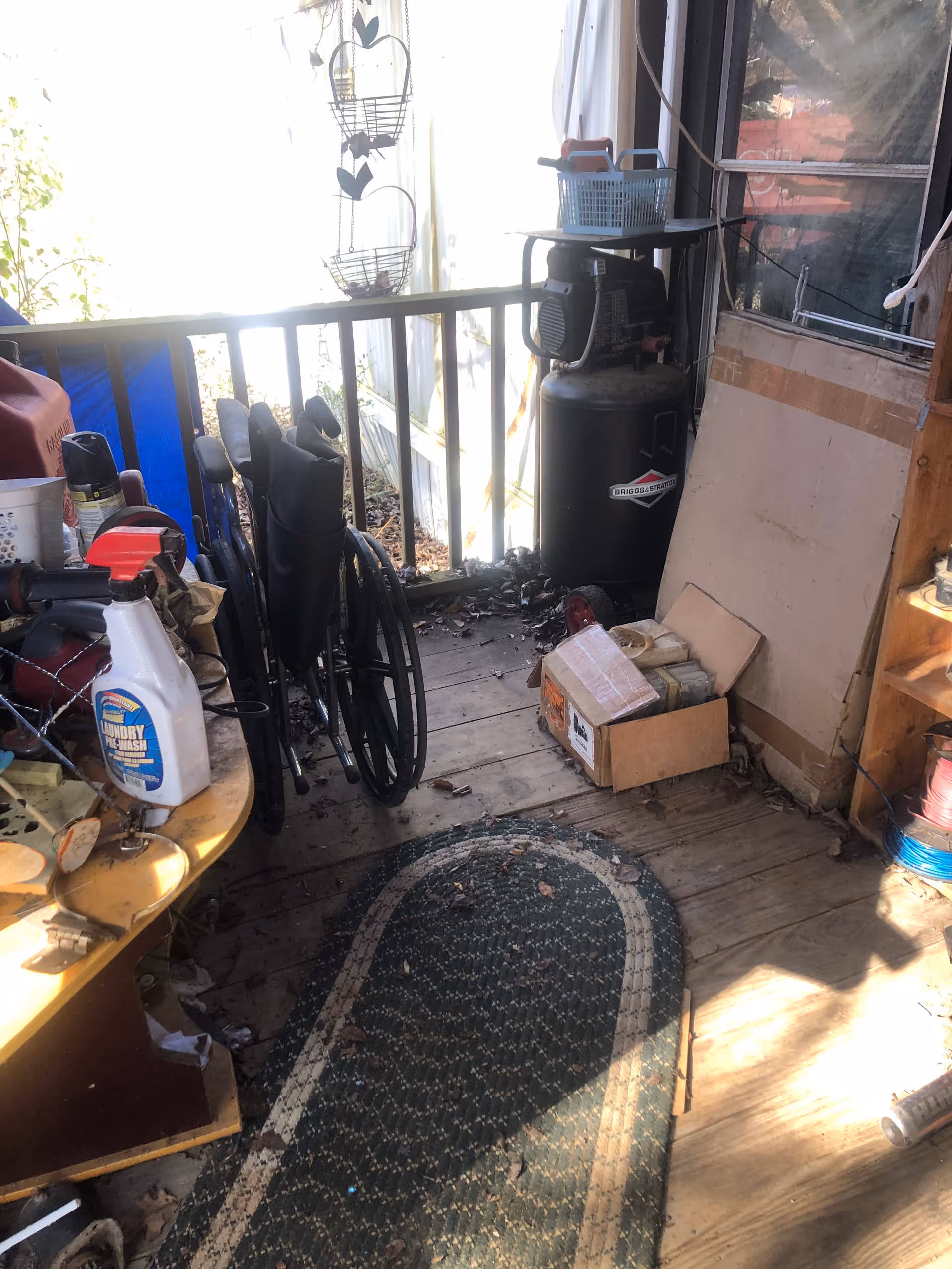 Cluttered outdoor porch with a wheelchair, cleaning supplies, boxes, and propane tank.
