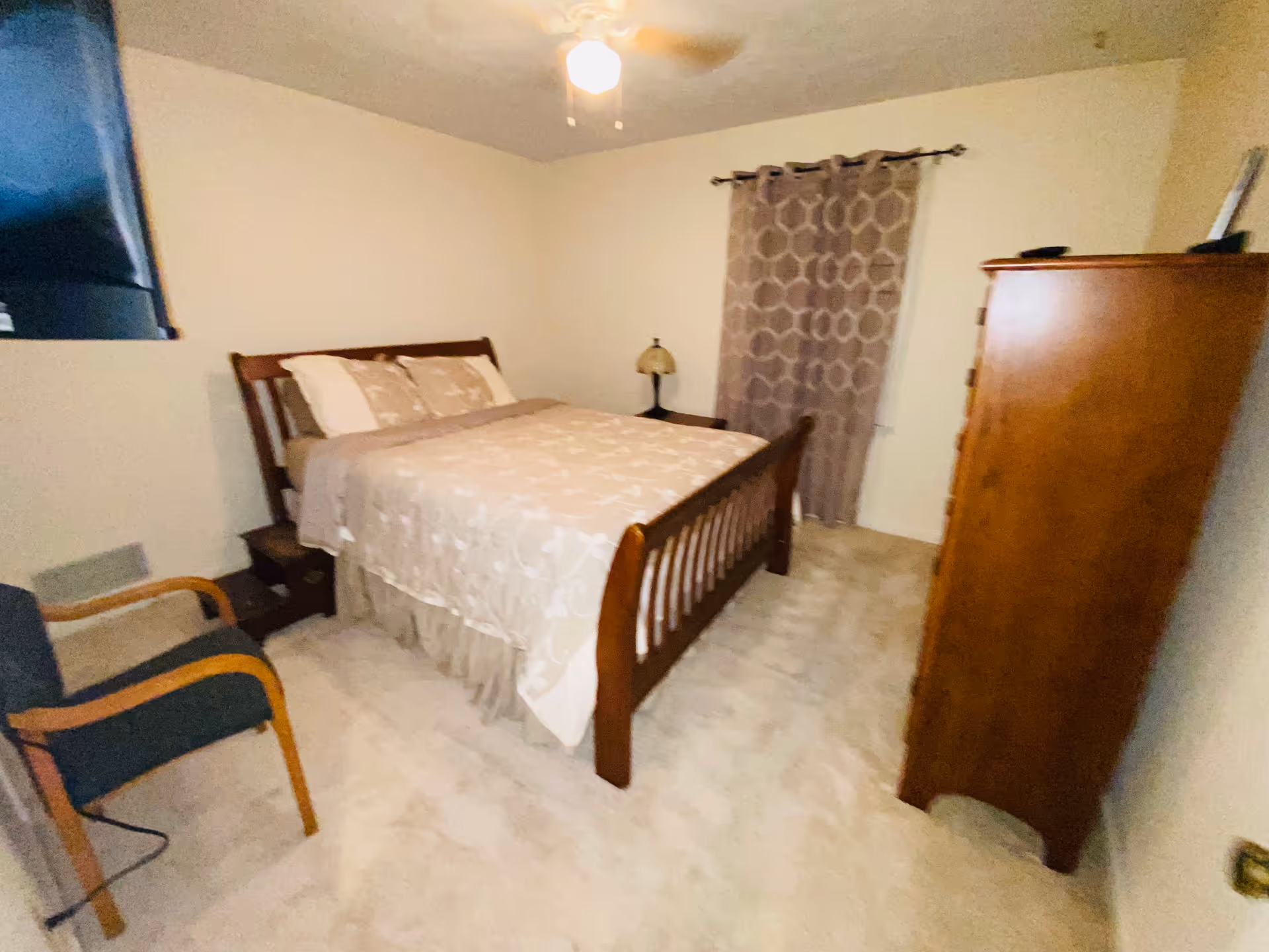 A cozy bedroom in a senior home with a bed, dresser, chair, and window curtain.