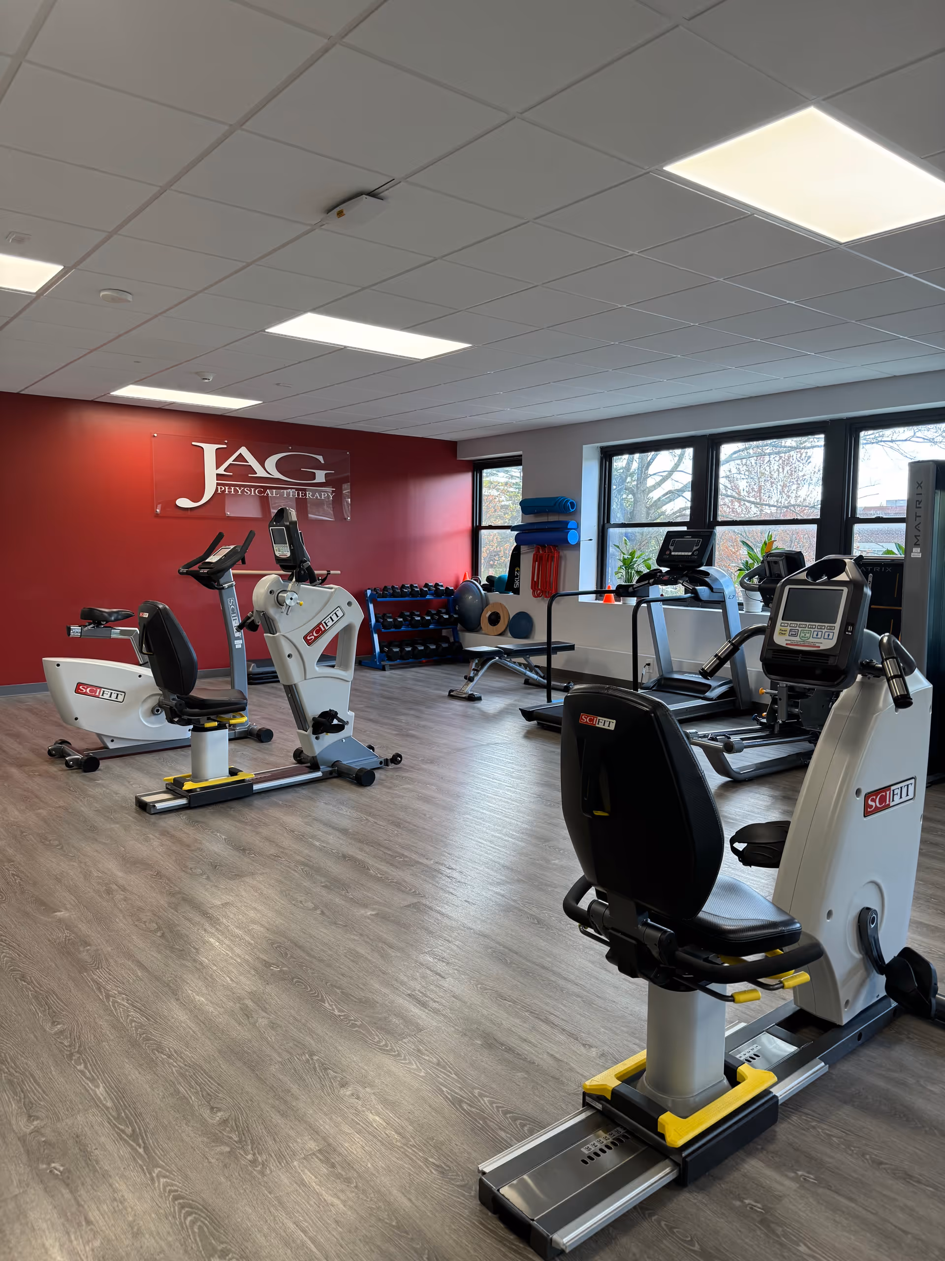 Interior fitness room at JAG Physical Therapy with cardio machines, weights, and a red accent wall.