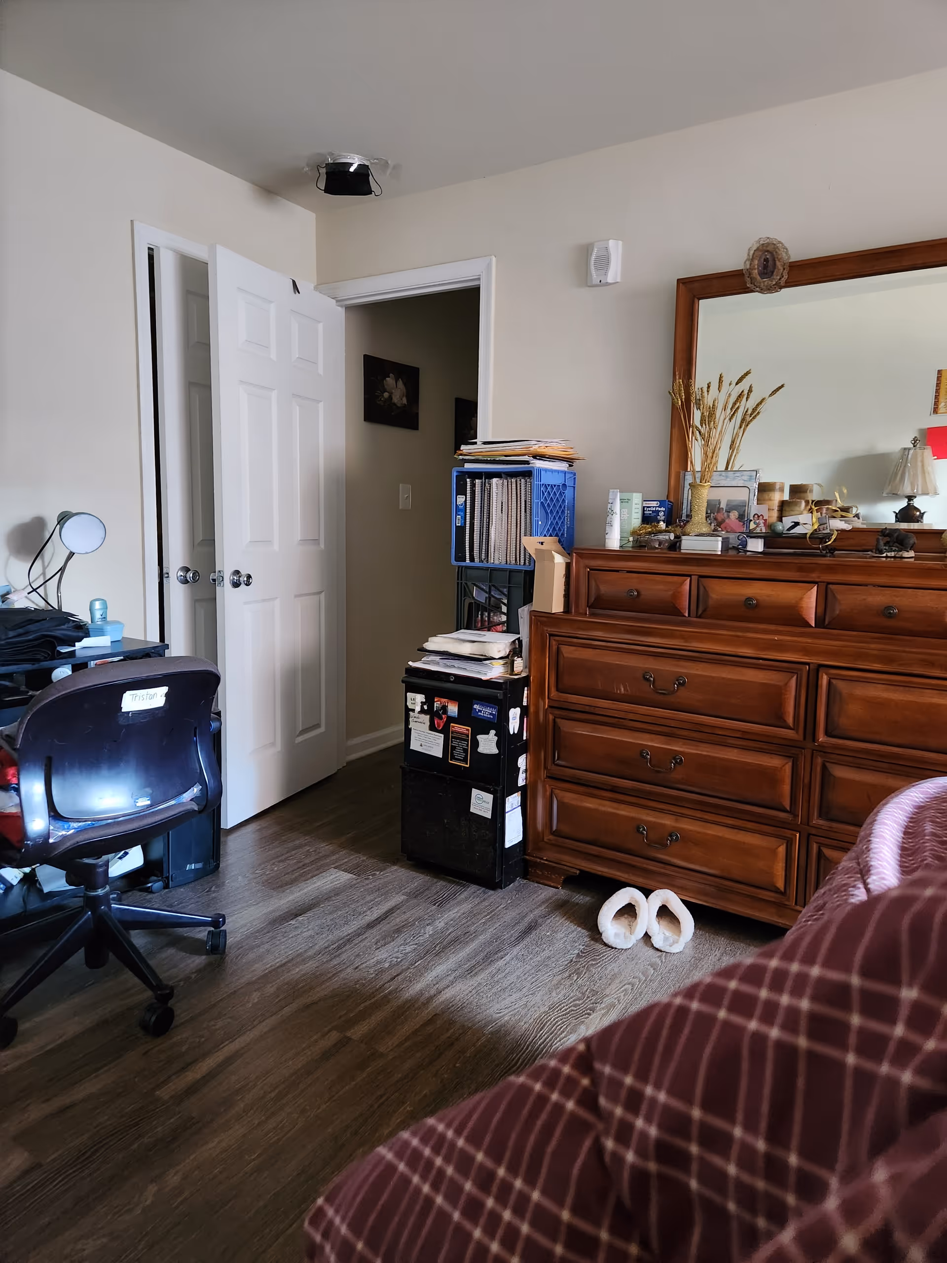 A cluttered bedroom with a wooden dresser, large mirror, and open doorway.