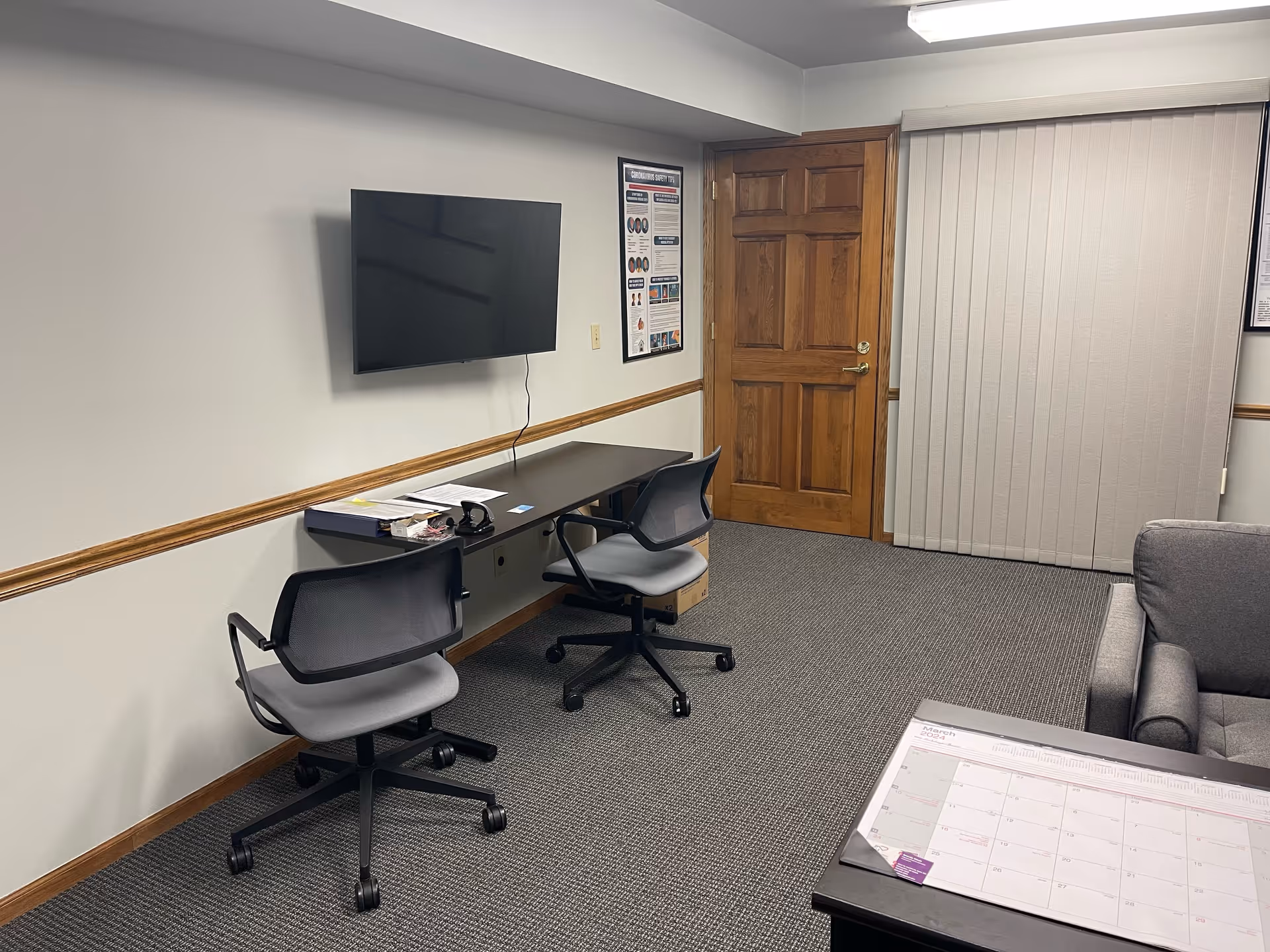A small office area with desks, chairs, a wall-mounted TV, and a wooden door.
