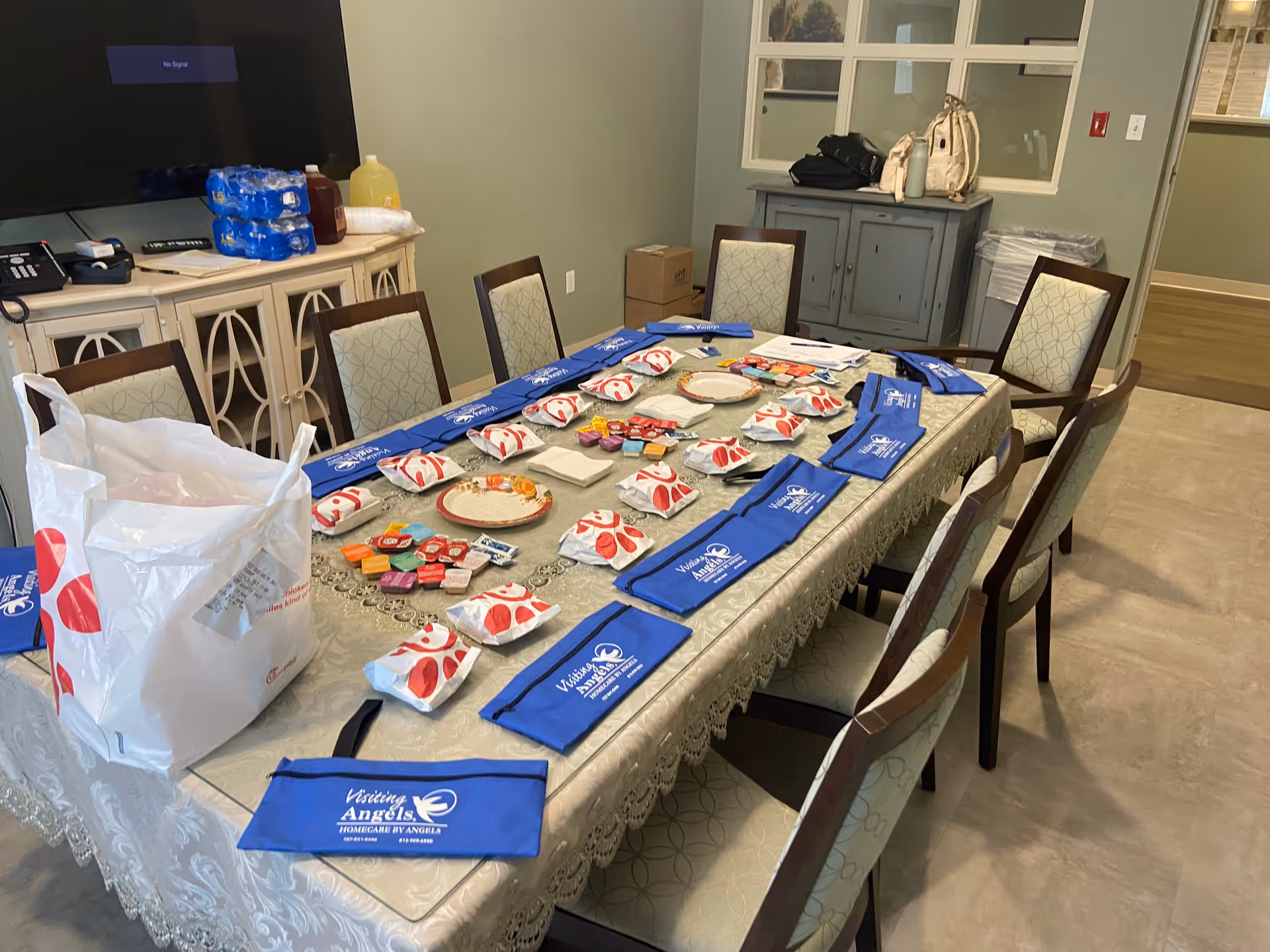 A long dining table is set with blue-branded napkins and assorted items in a care facility.