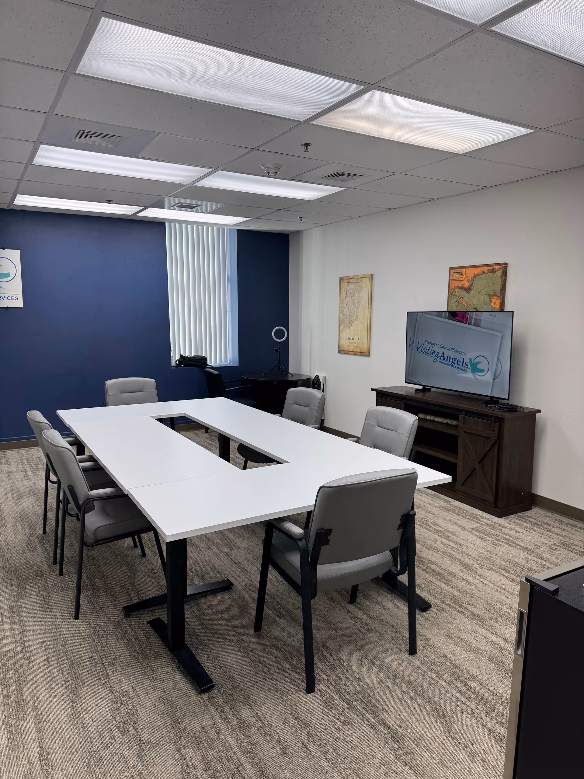 Conference room with white U-shaped table, gray chairs, and a TV displaying Visiting Angels.