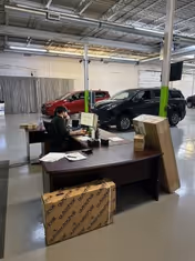 Office desk inside a showroom with a person, boxes, and two cars.