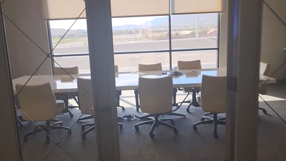 Interior conference room with white chairs around a long table and large windows.