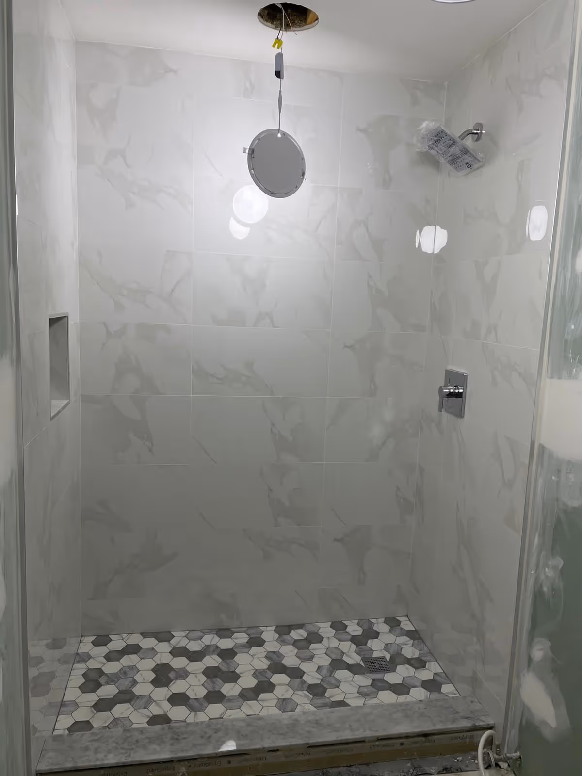 Shower stall with marble tile walls and hexagonal floor tiles, ceiling wiring exposed.